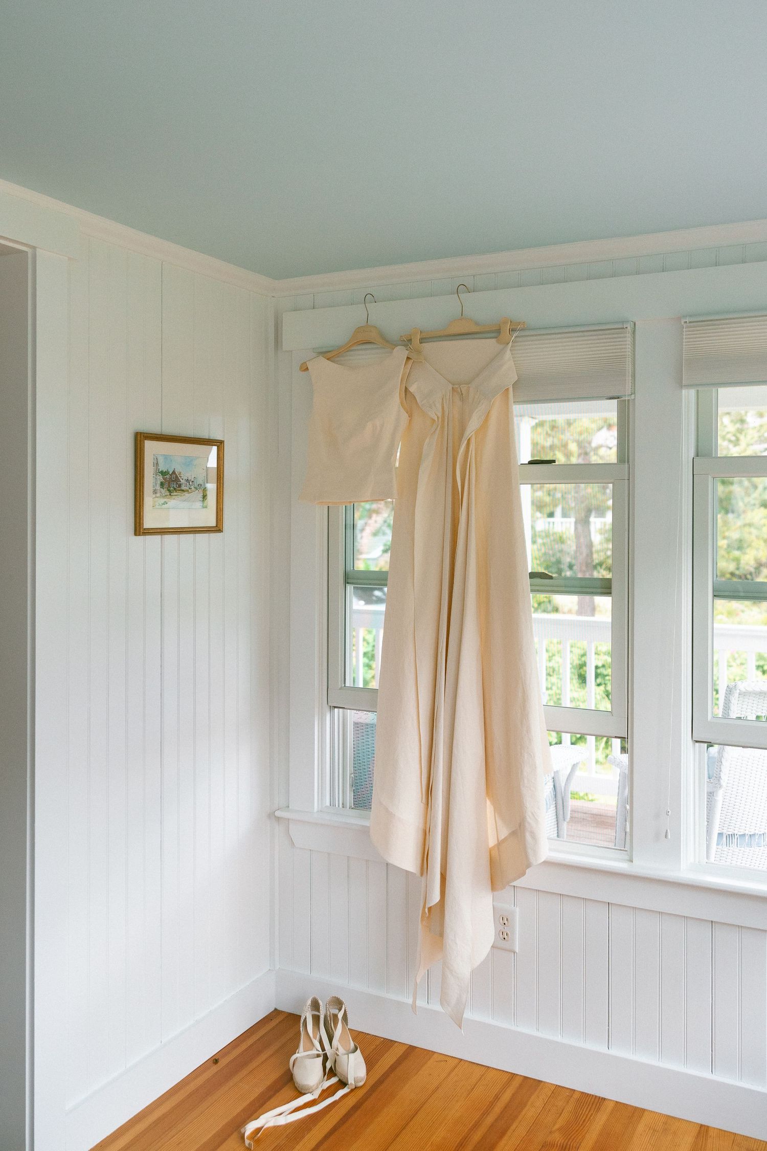 Elegant wedding dress hanging on wooden hangers by a sunlit window with heels on hardwood floor below.