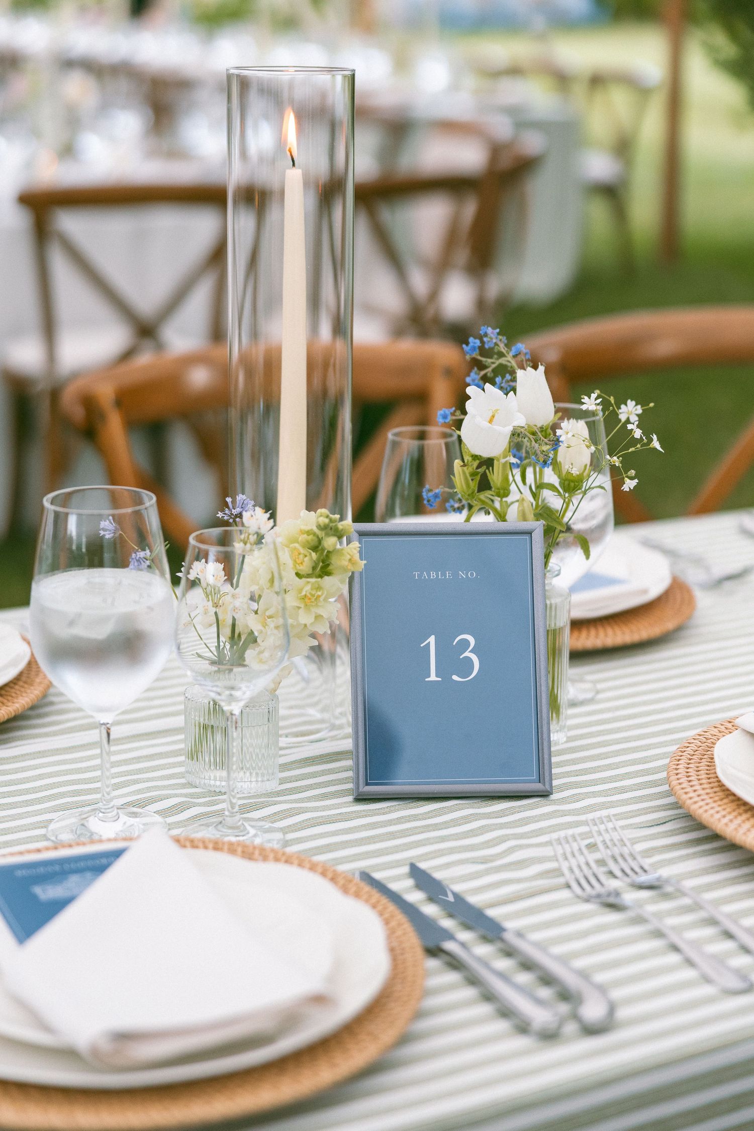 Elegant wedding table setting with blue table number 13, candle, floral arrangements, woven chargers, and striped linen.