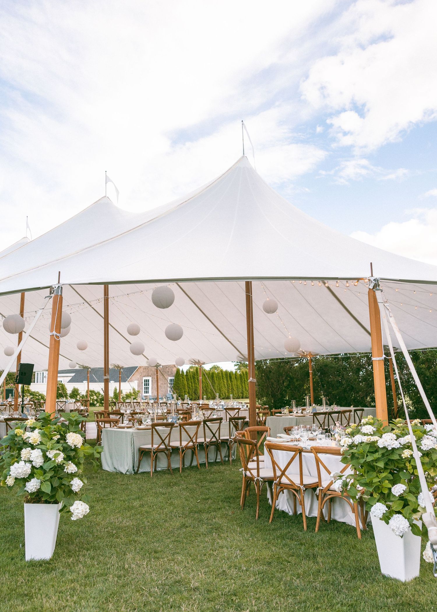 Elegant outdoor wedding reception under a white sailcloth tent with wood chairs, white floral arrangements, and gray lanterns.