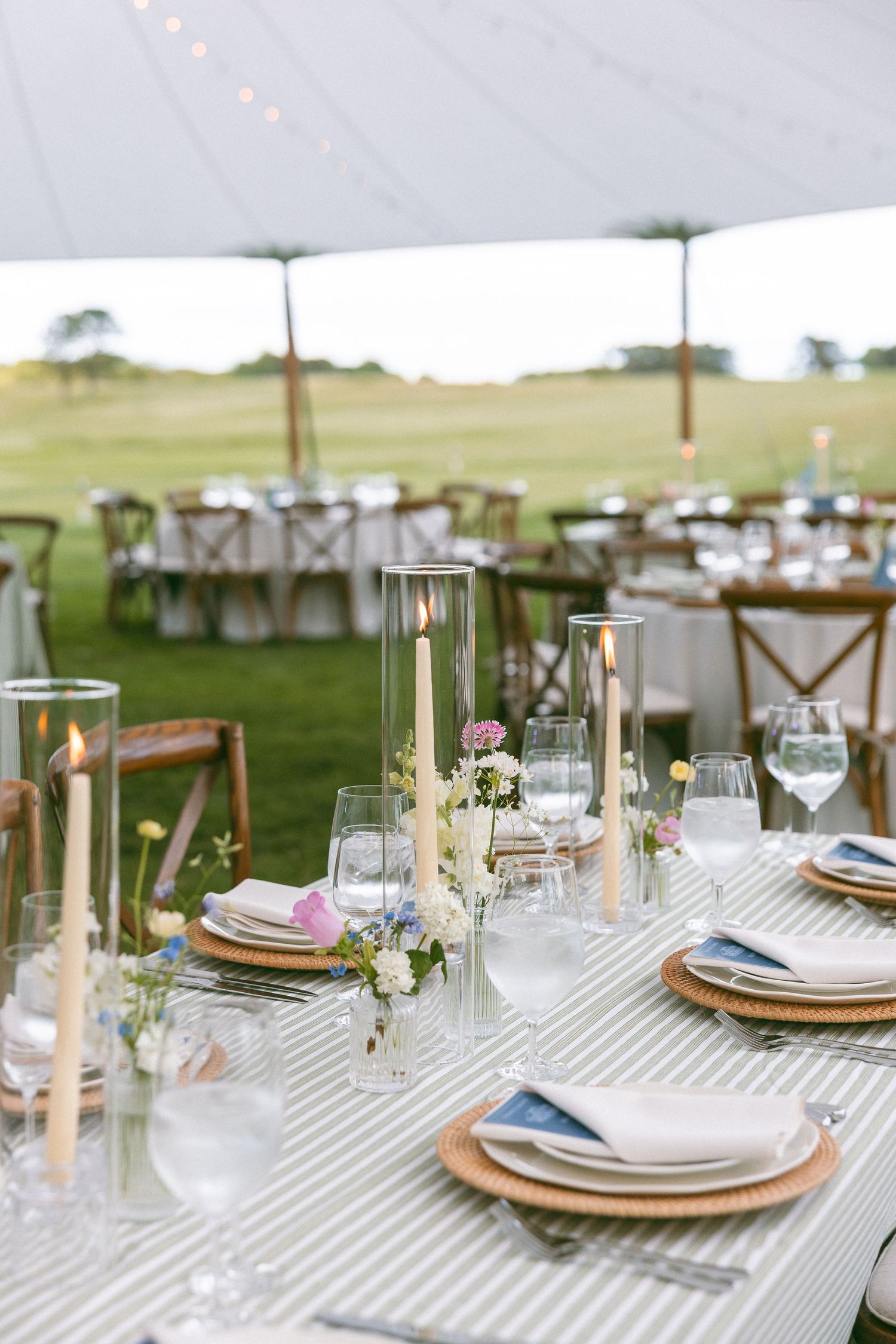 Elegant outdoor wedding reception table setting with candles, floral arrangements, striped linens, and glassware under a white tent.