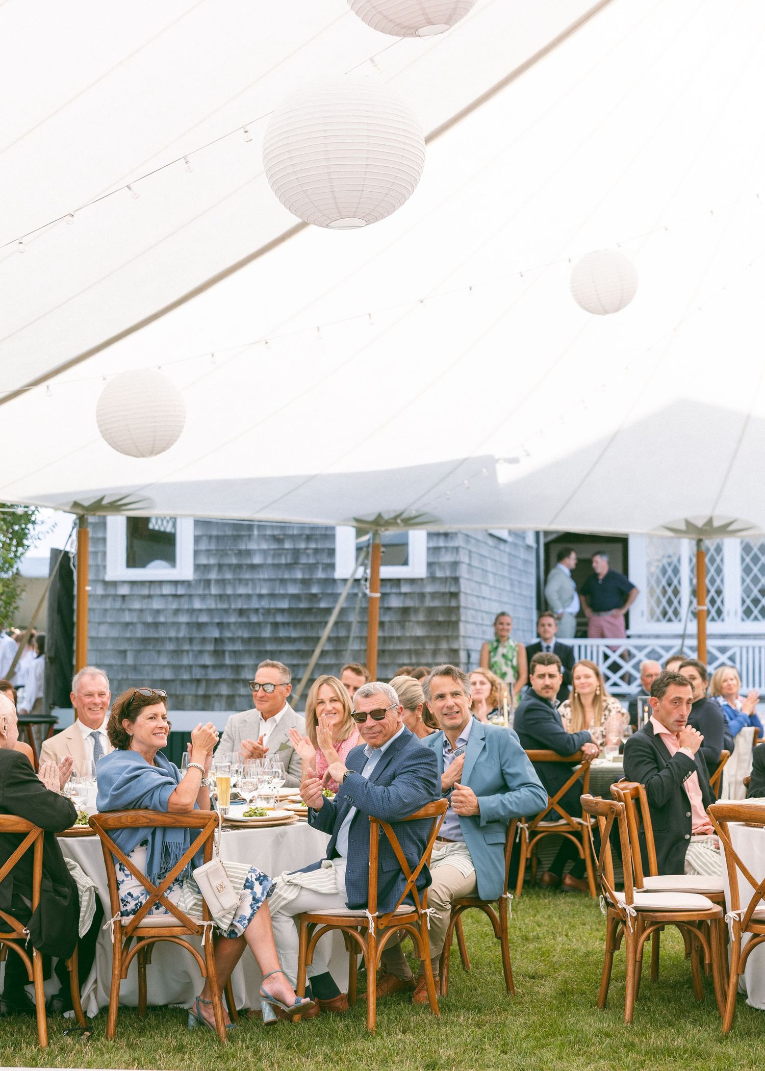 Guests applaud at an outdoor wedding reception under a white tent with paper lantern decorations on a sunny day.