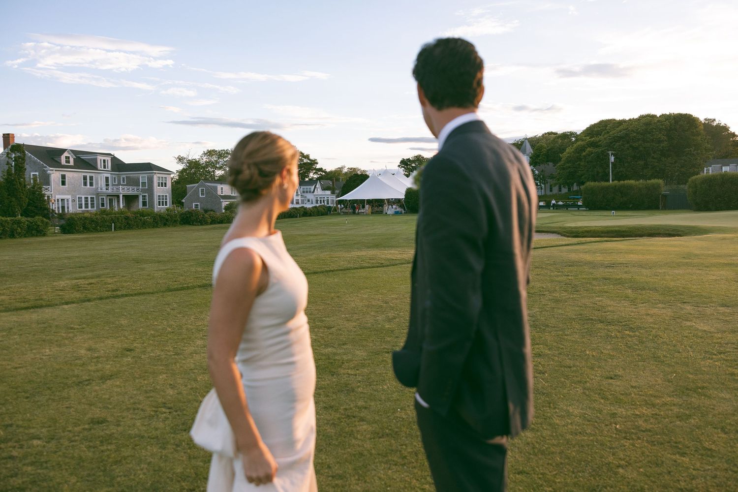 A bride and groom stand on a green lawn at sunset, gazing toward a elegant estate venue.