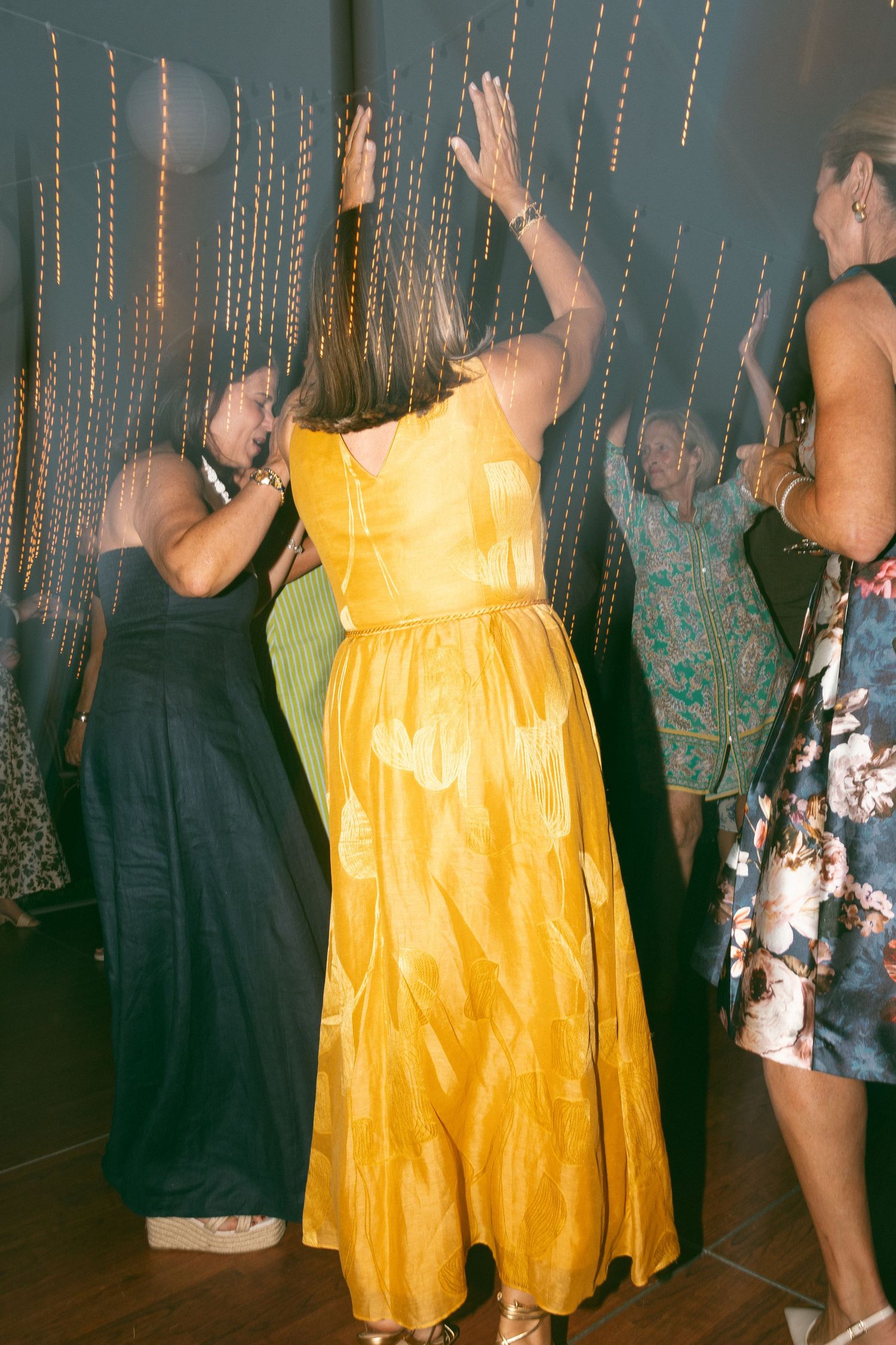 Women in elegant dresses dancing with hands raised at a formal evening event with golden curtain backdrop.
