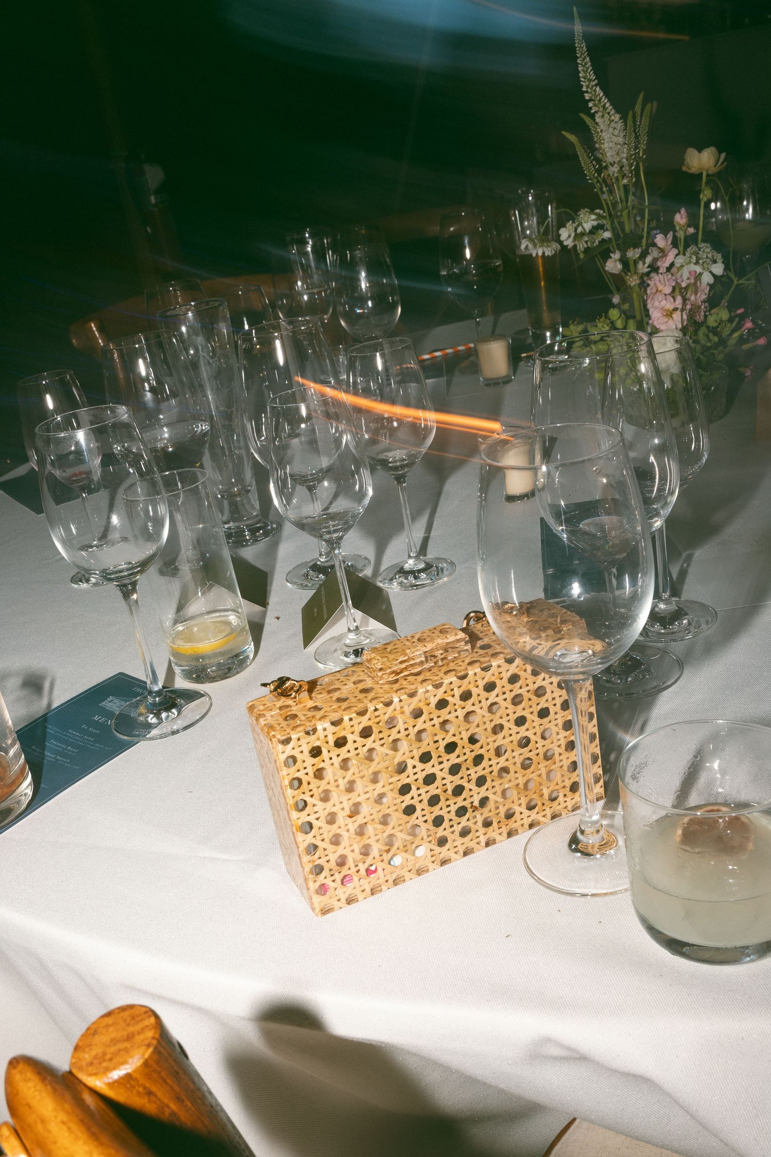Elegant wedding reception table with crystal glasses, floral centerpiece, and a woven gold clutch purse on white linen.