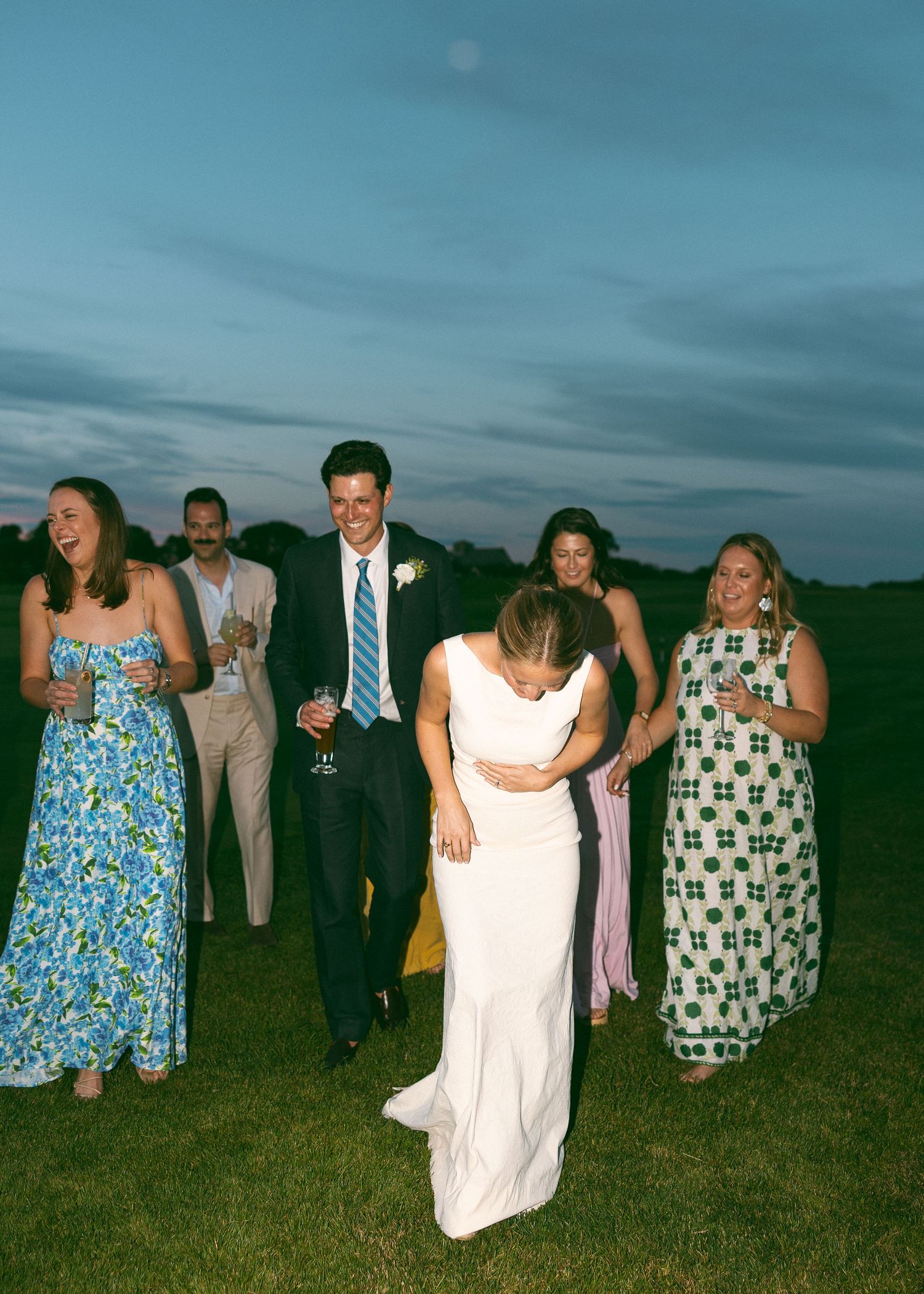 Bride and groom celebrate outdoors at dusk with guests in colorful dresses during wedding reception.