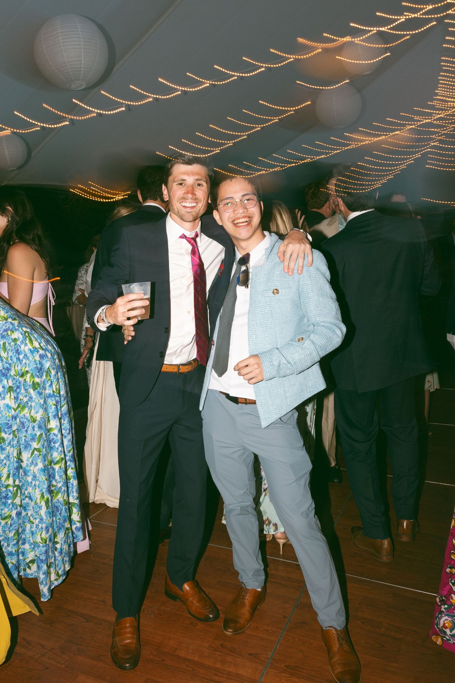 Two men in suits smiling and posing together at an elegant wedding reception with string lights.
