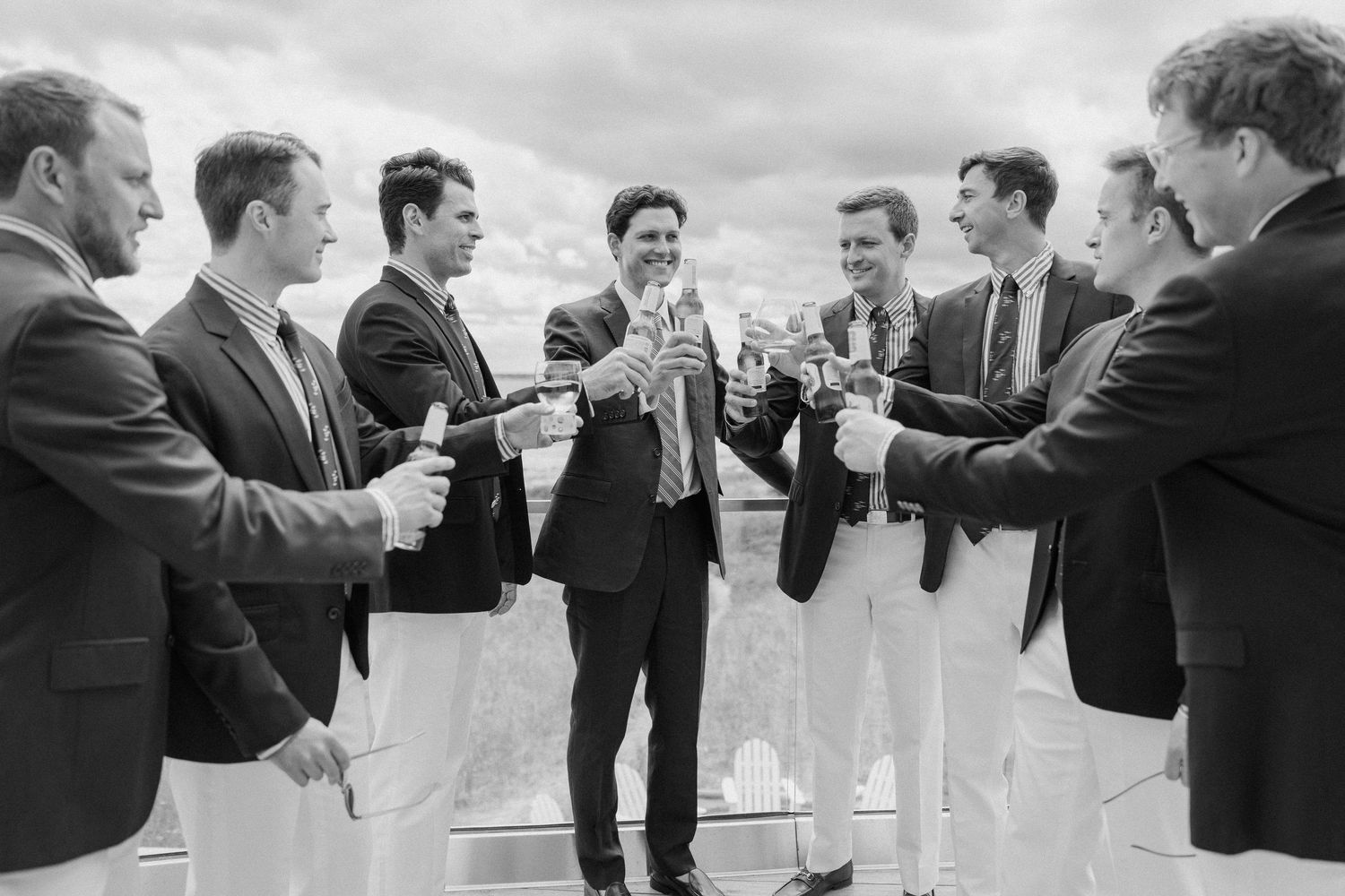 Black and white photo of groomsmen in suits toasting with champagne glasses outdoors at a wedding celebration.