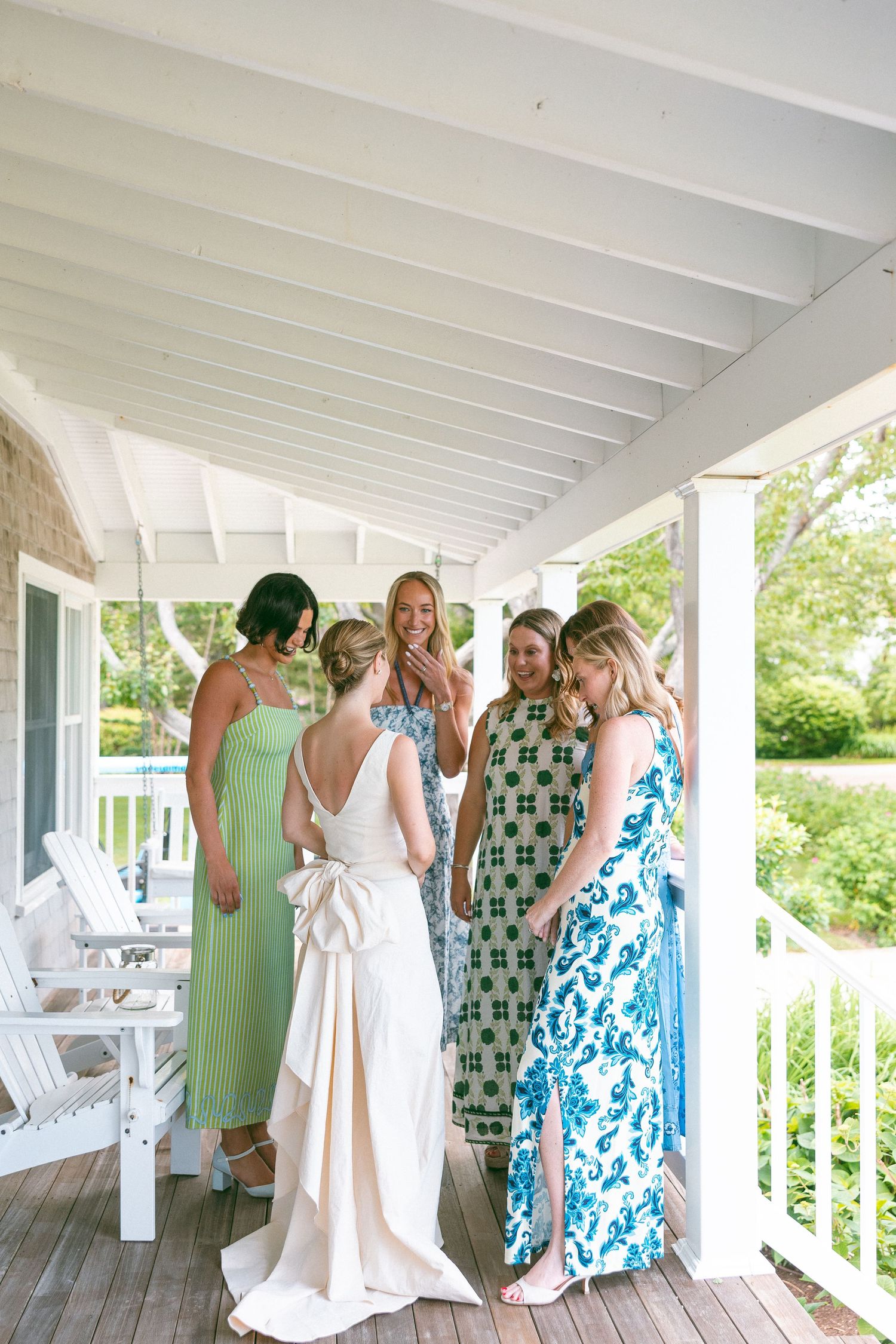 Bride in white gown with bow back shares a joyful moment with bridesmaids in green and blue dresses on a white porch.