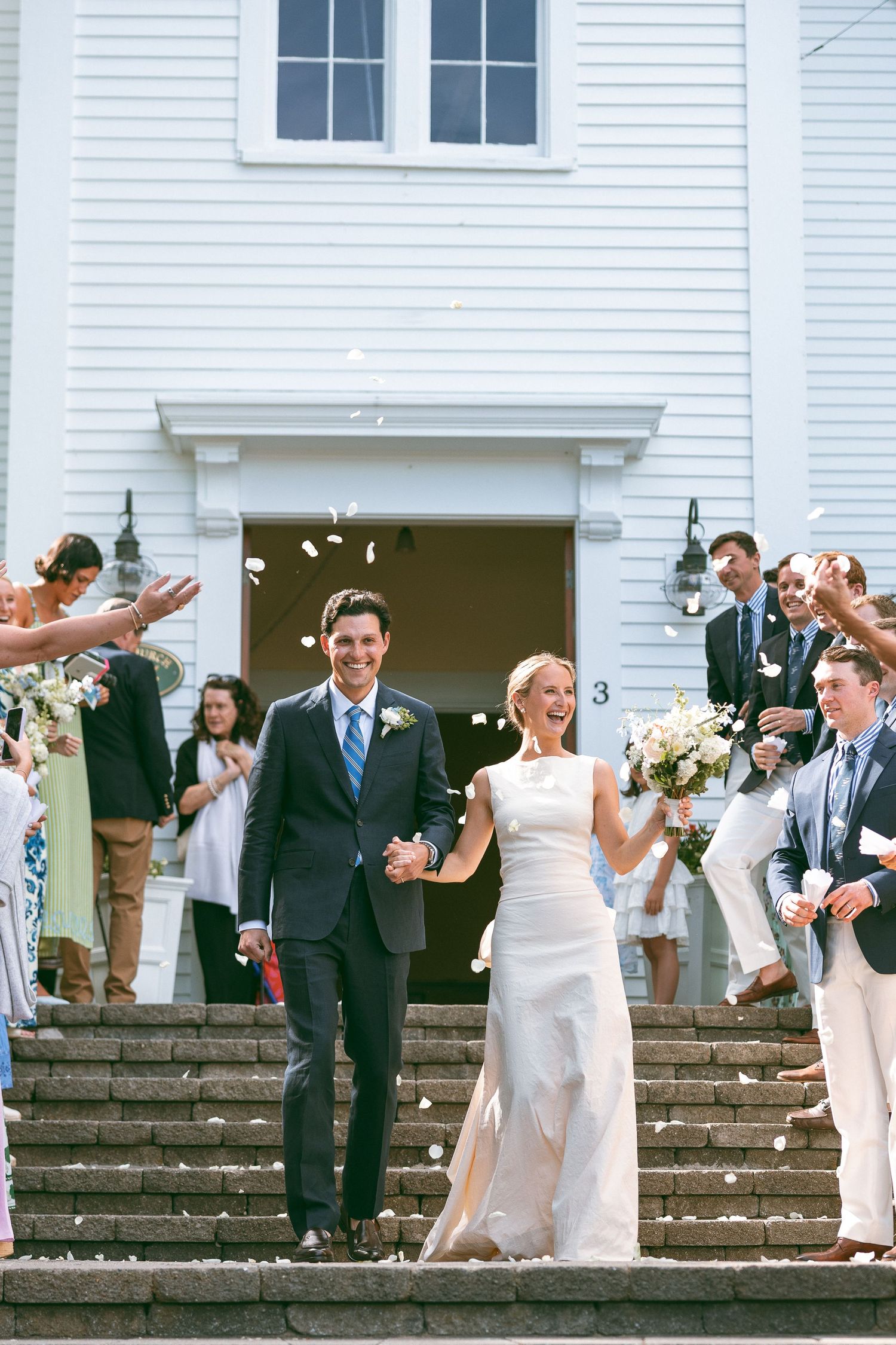 Newlywed couple exits white church steps as guests toss flower petals in joyful wedding ceremony sendoff.