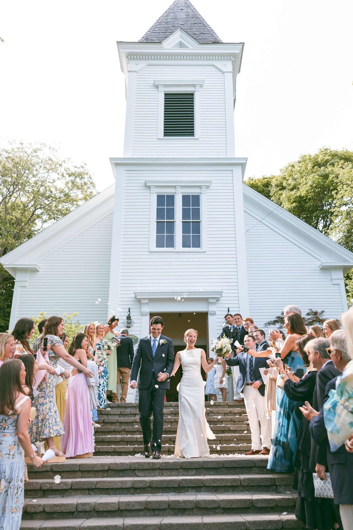 Bride and groom exit white New England chapel on stone steps, greeted by cheering wedding guests throwing petals.