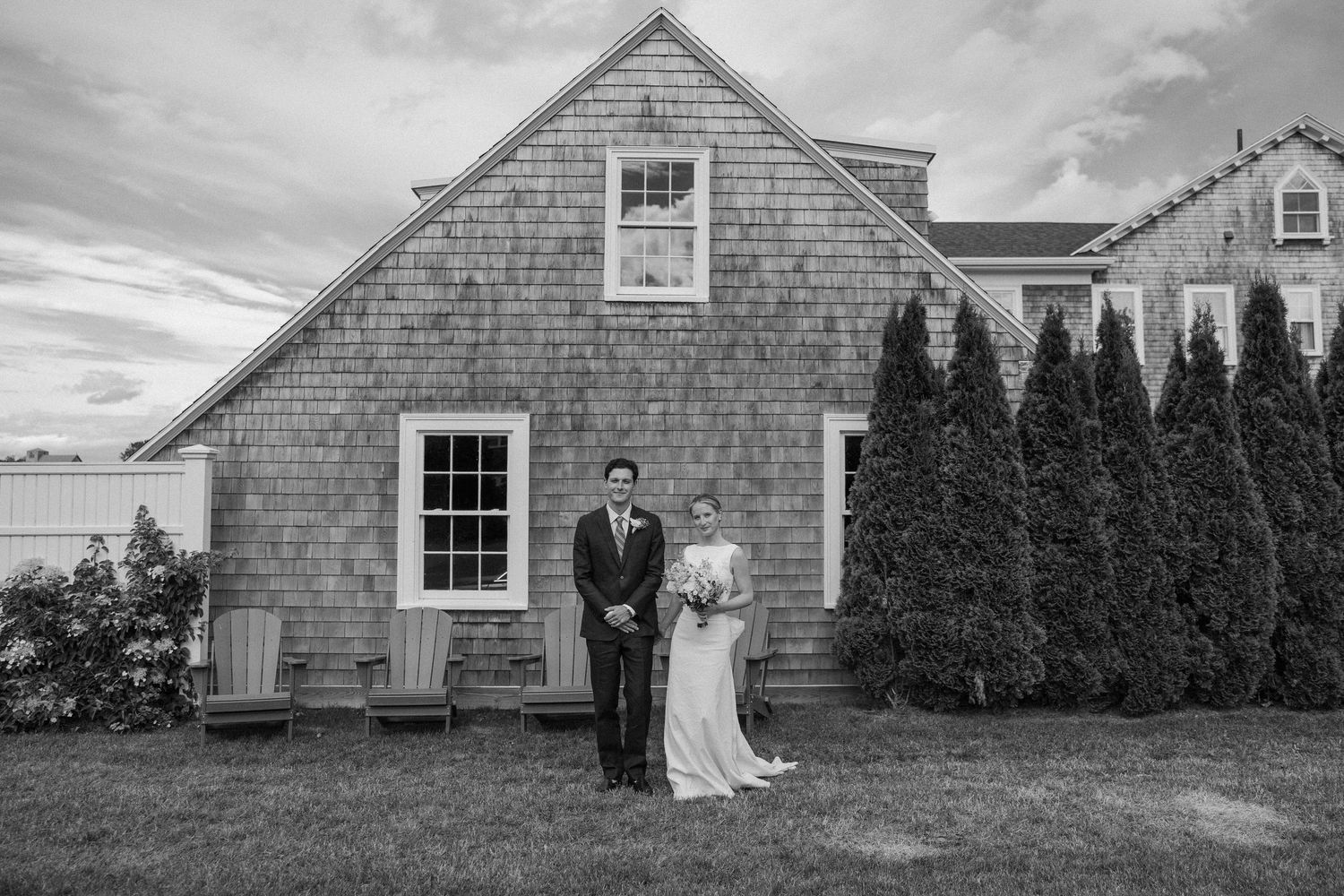 Black and white photo of a bride and groom standing outside a rustic shingle-style home with evergreen trees.