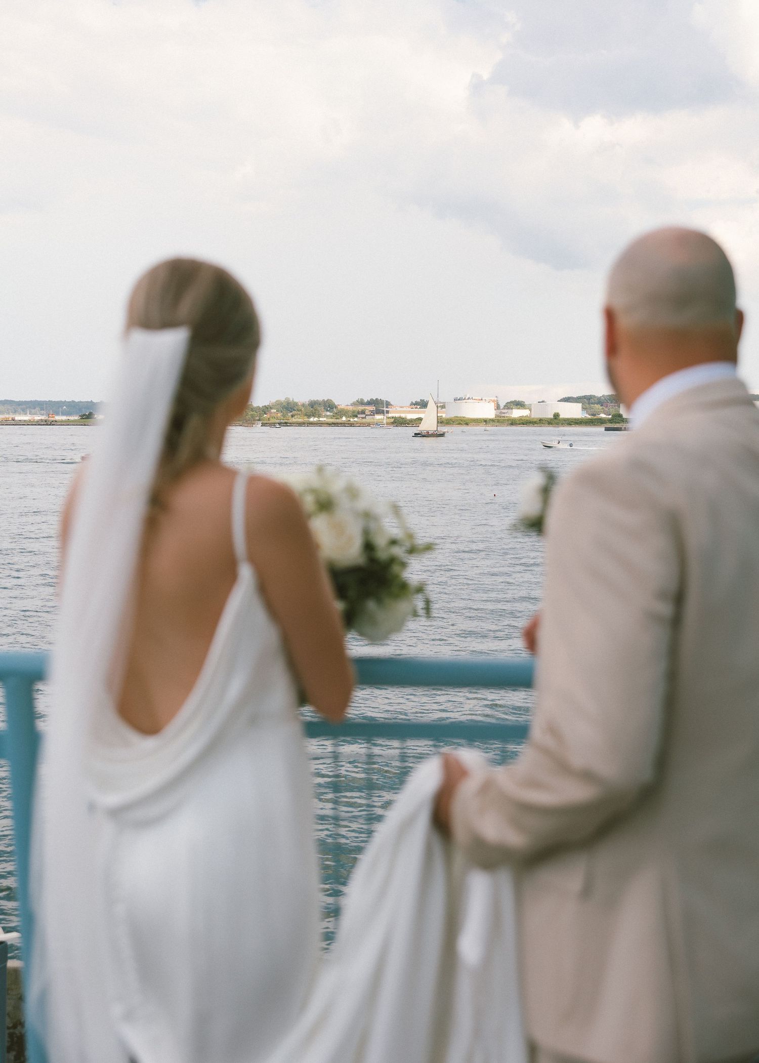 A couple stands together against a waterfront railing with boats visible in the background.