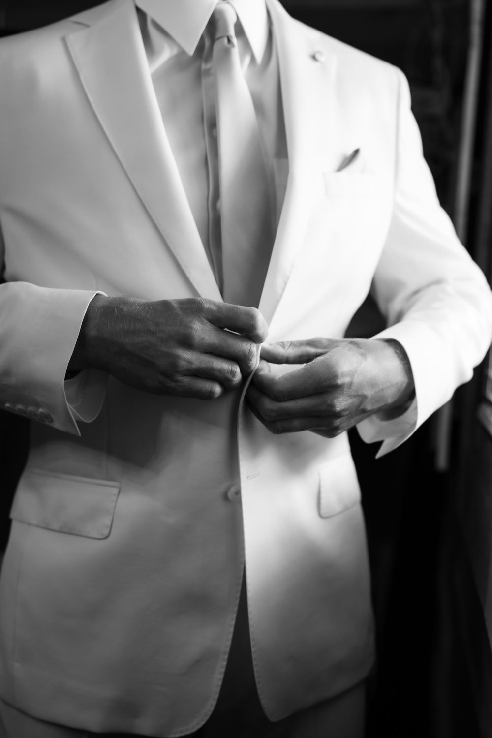Black and white photo of a groom in a white suit buttoning his jacket before his wedding ceremony.