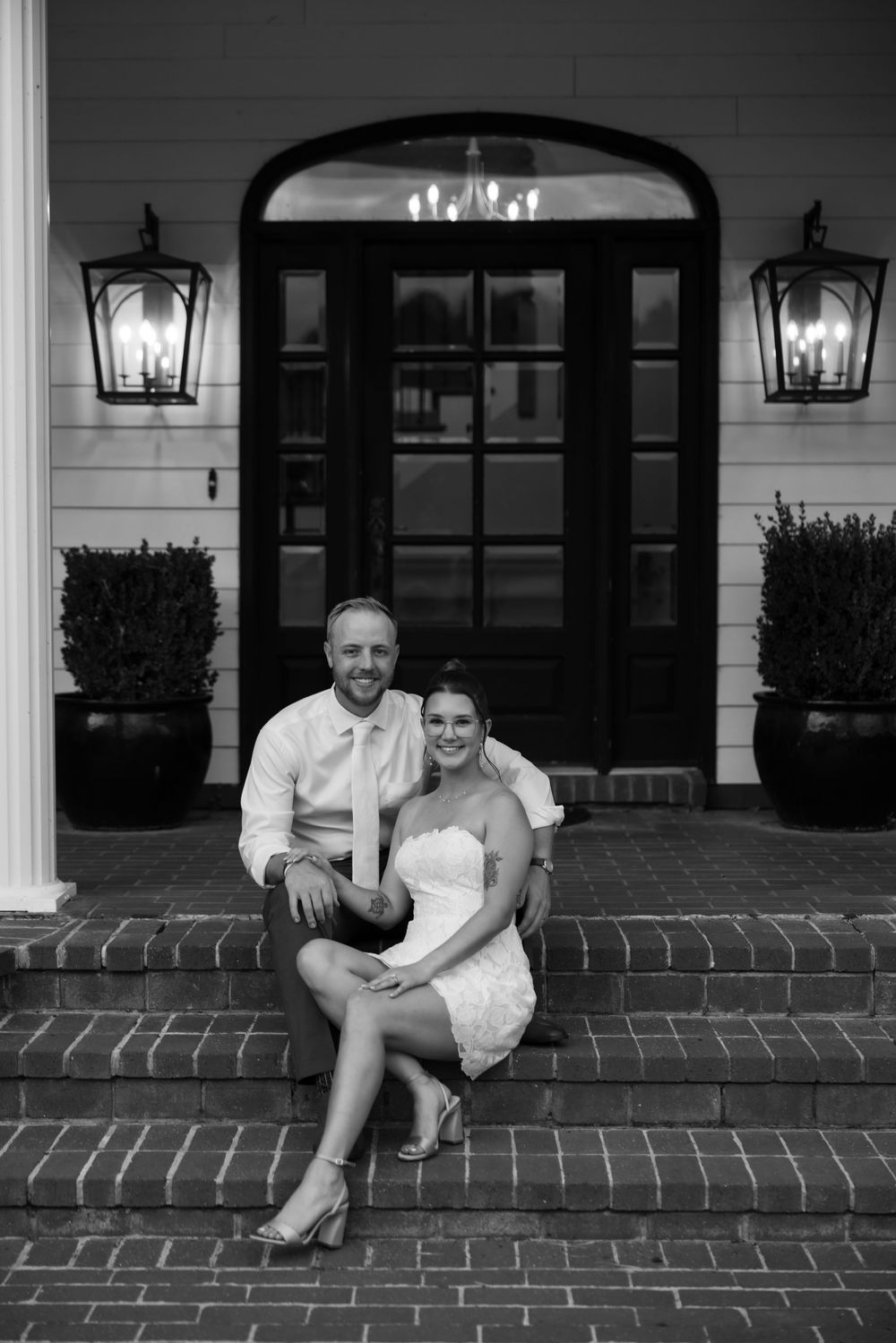 Couple in wedding attire sits on brick steps outside elegant venue in black and white portrait photo.