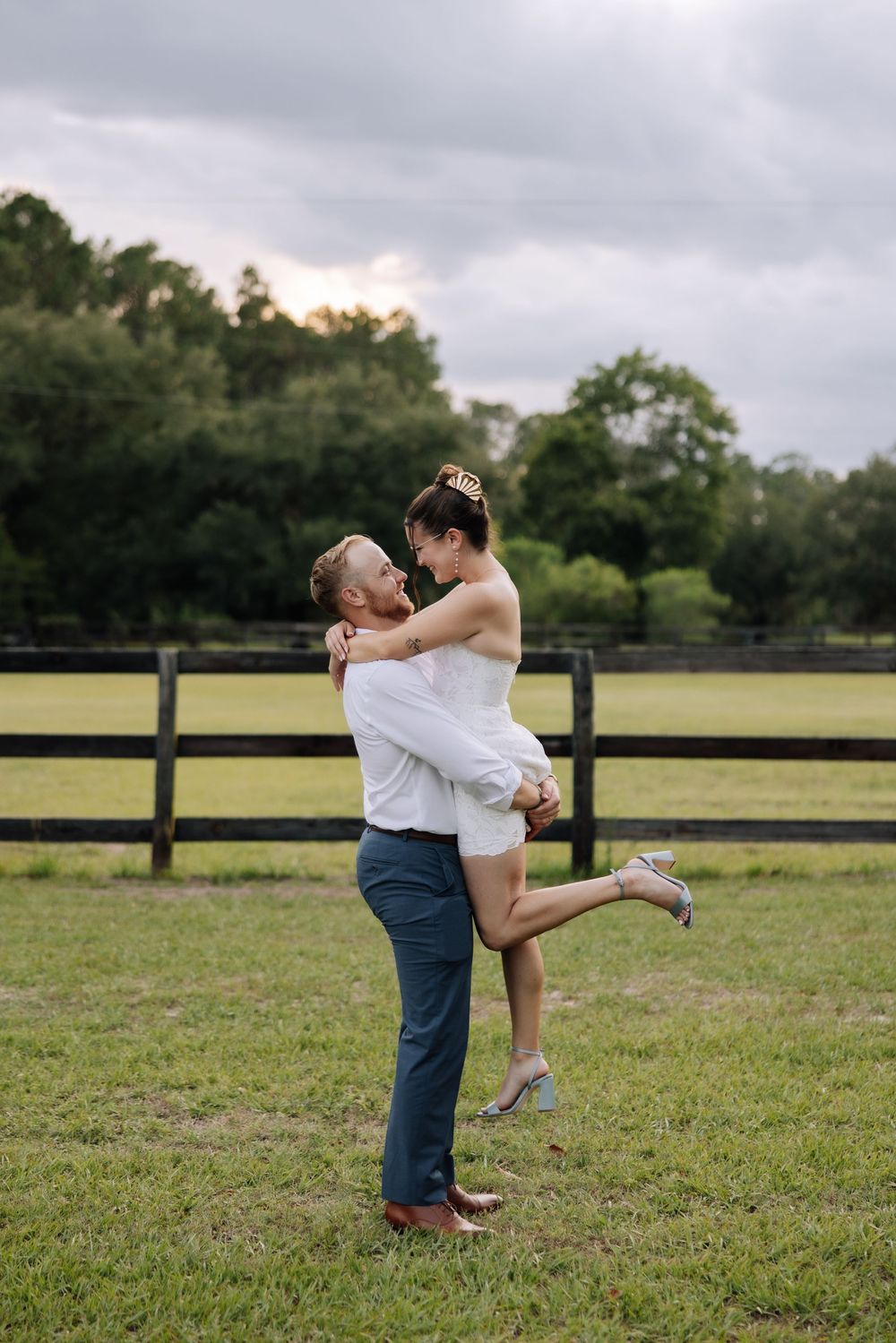 Groom lifts bride in white dress at rustic farm wedding venue with wooden fence and green fields.