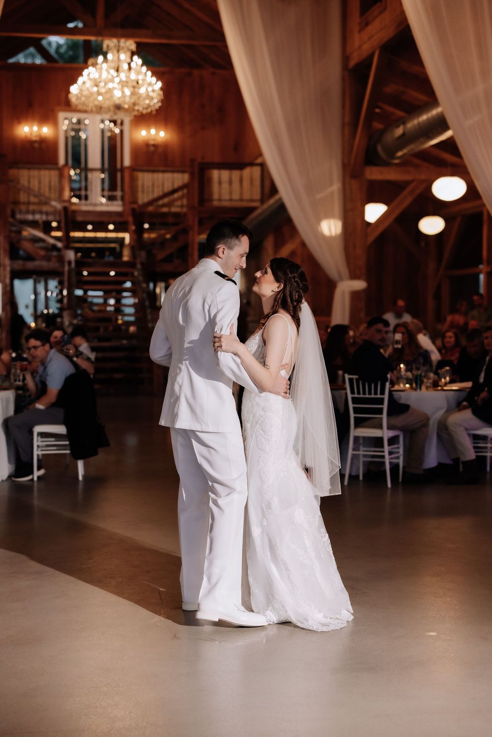 Bride and groom share their first dance in a rustic barn wedding venue with warm lighting and chandelier.