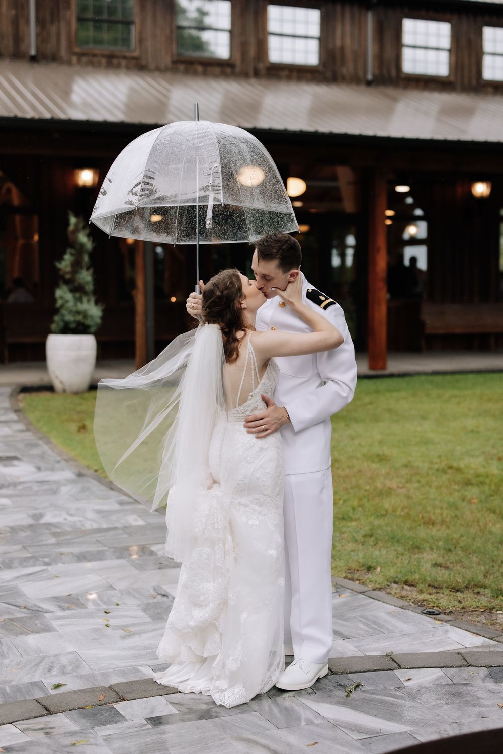 Bride and groom kissing under a clear umbrella in the rain, bride in lace gown with veil, groom in white military uniform.