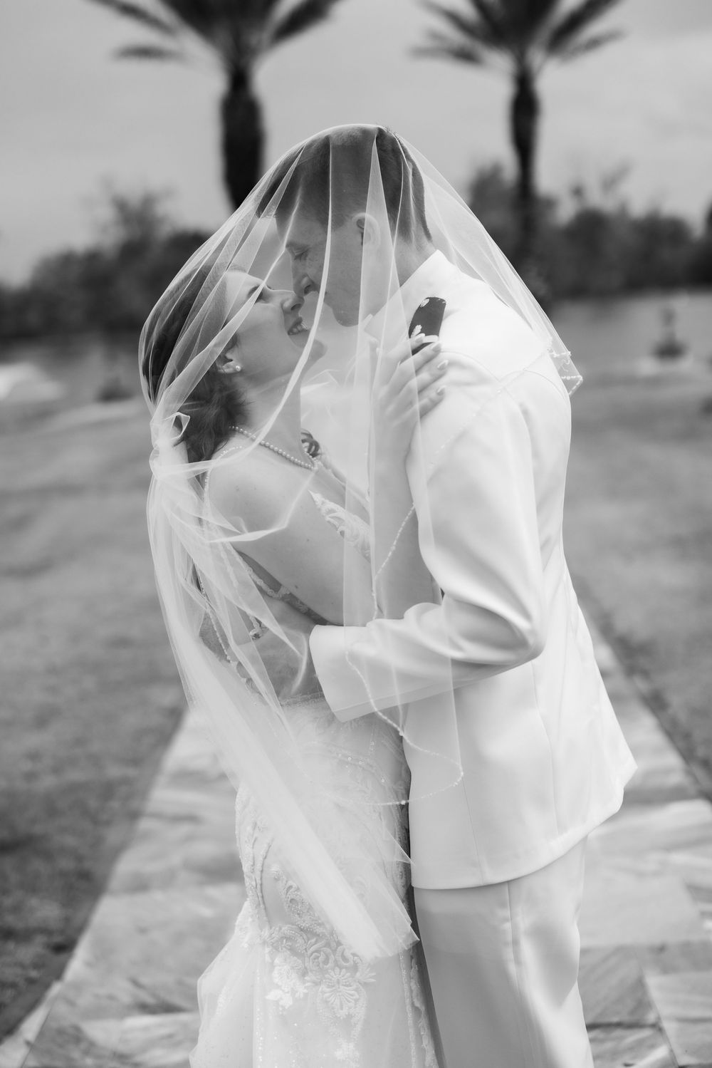 Black and white photo of bride and groom sharing a kiss under a flowing veil outdoors on their wedding day.