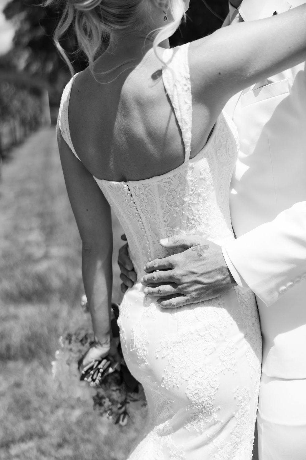Black and white photo of bride in lace wedding gown with groom's hand on her waist outdoors.
