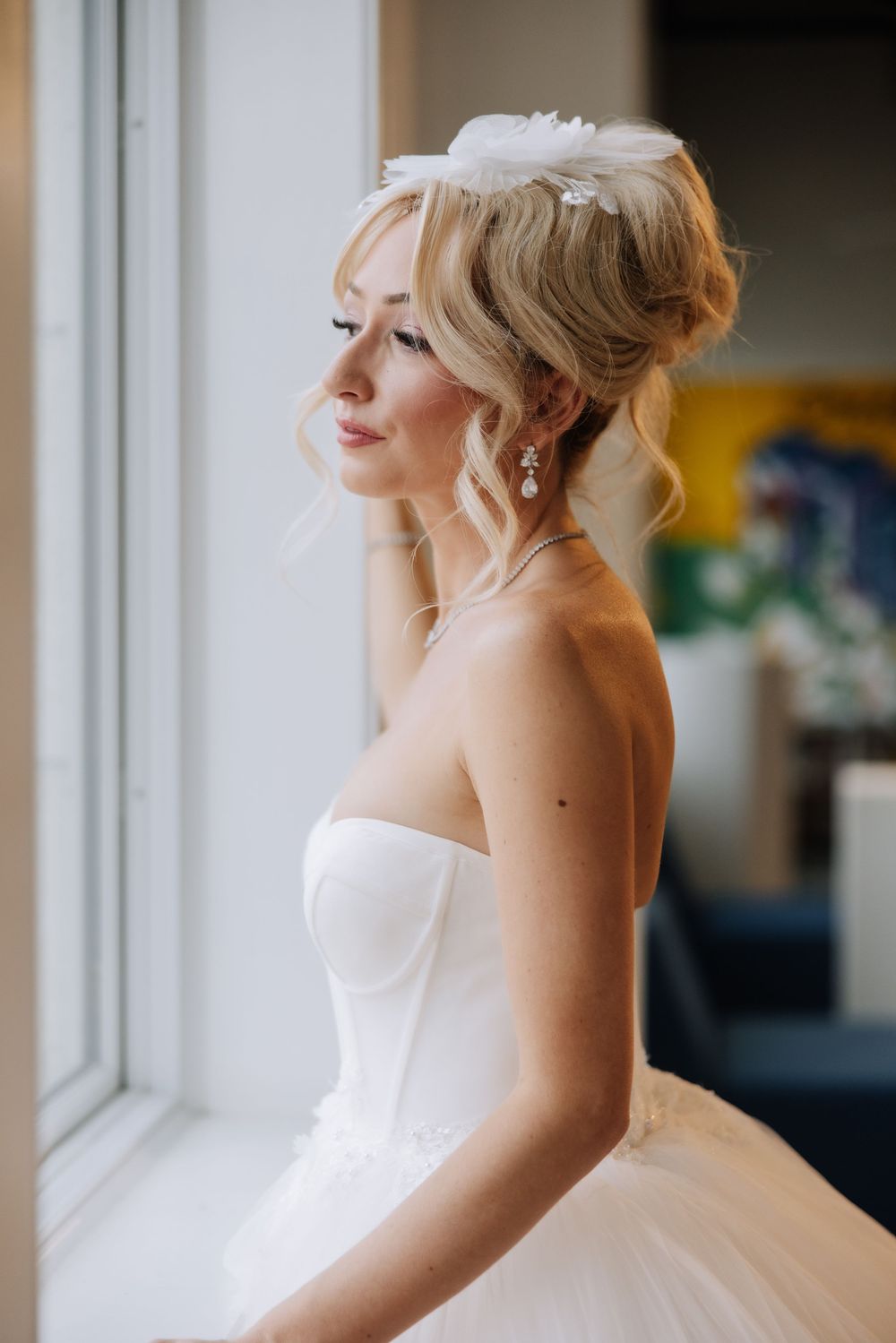 Elegant bride in strapless white gown with floral hair accessory gazing out window in soft natural light.