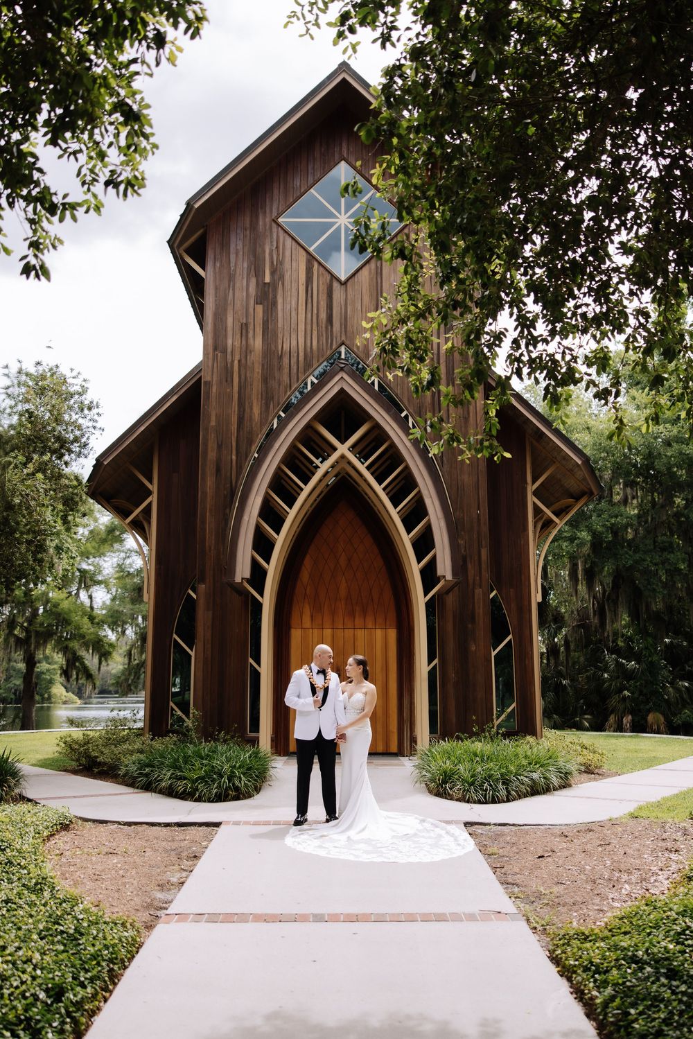 Bride and groom stand before a stunning rustic wooden chapel with Gothic arched doors, surrounded by lush greenery.