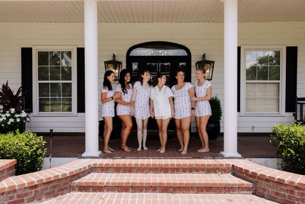 Six bridesmaids in matching striped rompers pose together on a white Southern-style home's brick front porch steps.