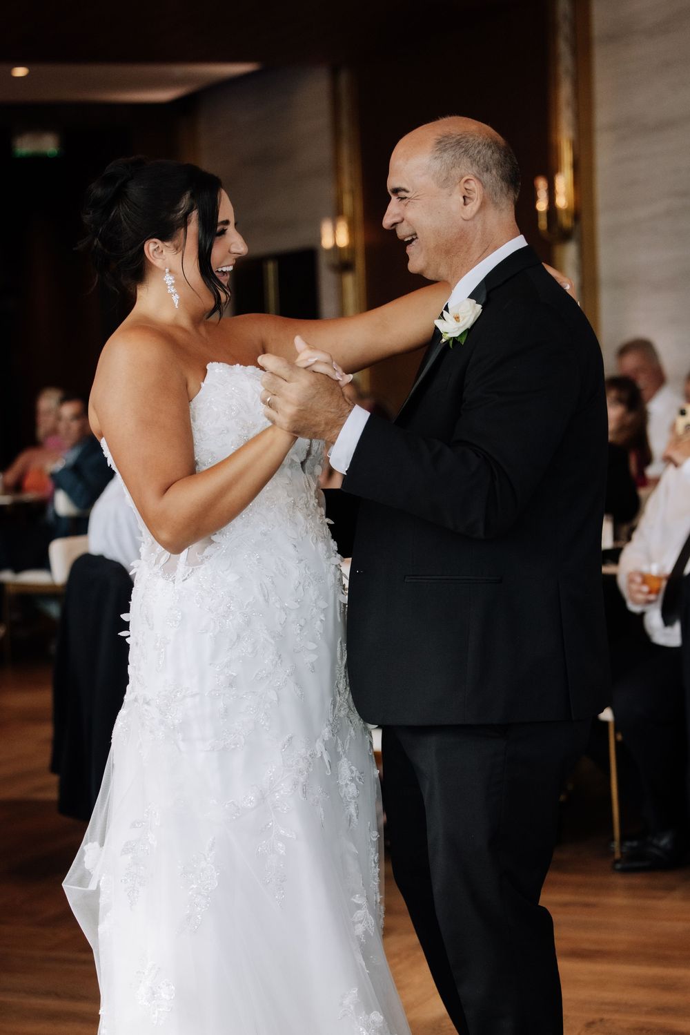 Bride in white lace gown shares a joyful first dance with her father in a black tuxedo at wedding reception.