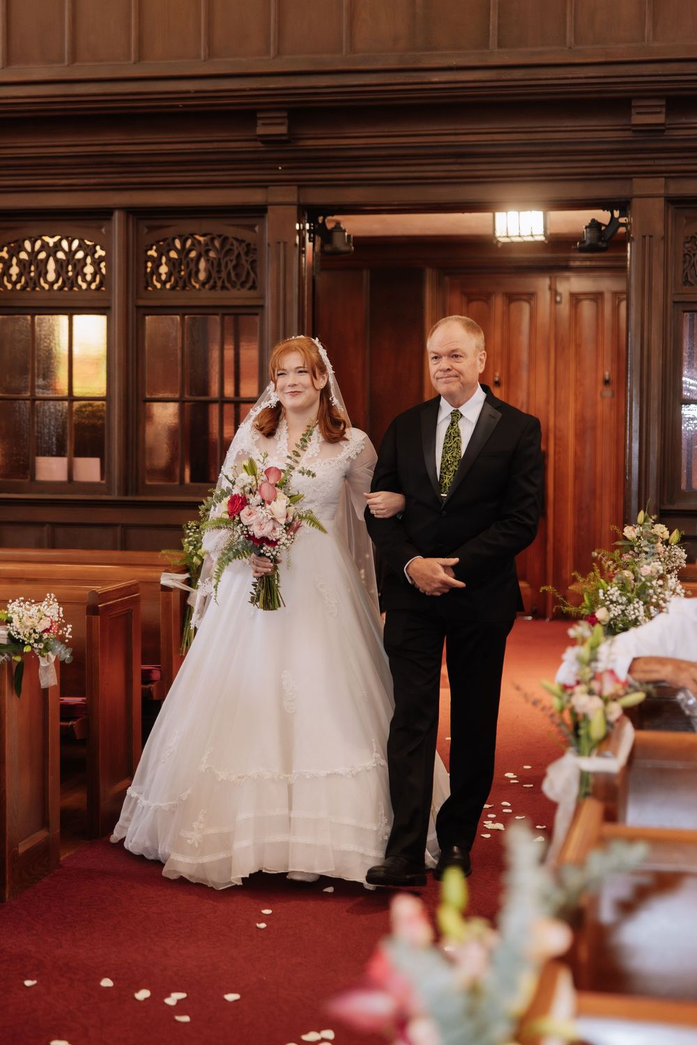 Bride in white gown walks down church aisle with father, holding pink bouquet, rose petals on red carpet.