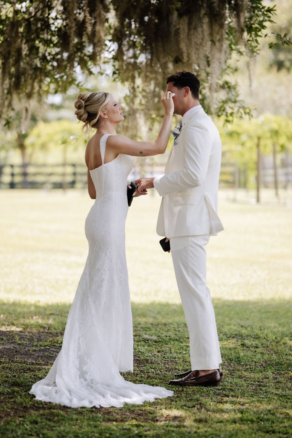 Bride in white gown gently touches groom's face as they share a tender moment outdoors under moss-draped trees.