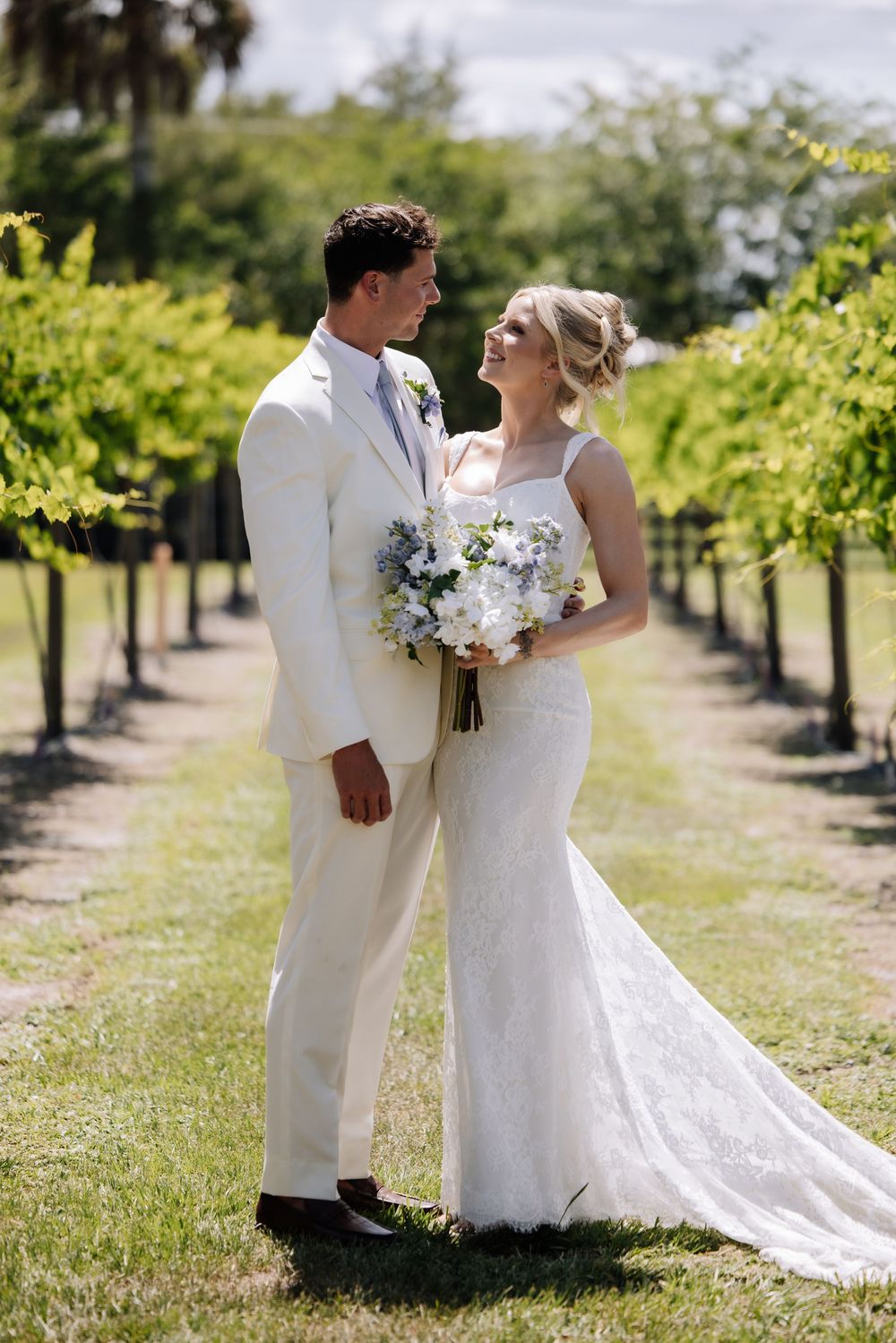 Bride and groom share a joyful moment in a vineyard, she holds white blooms, both dressed in elegant wedding attire.