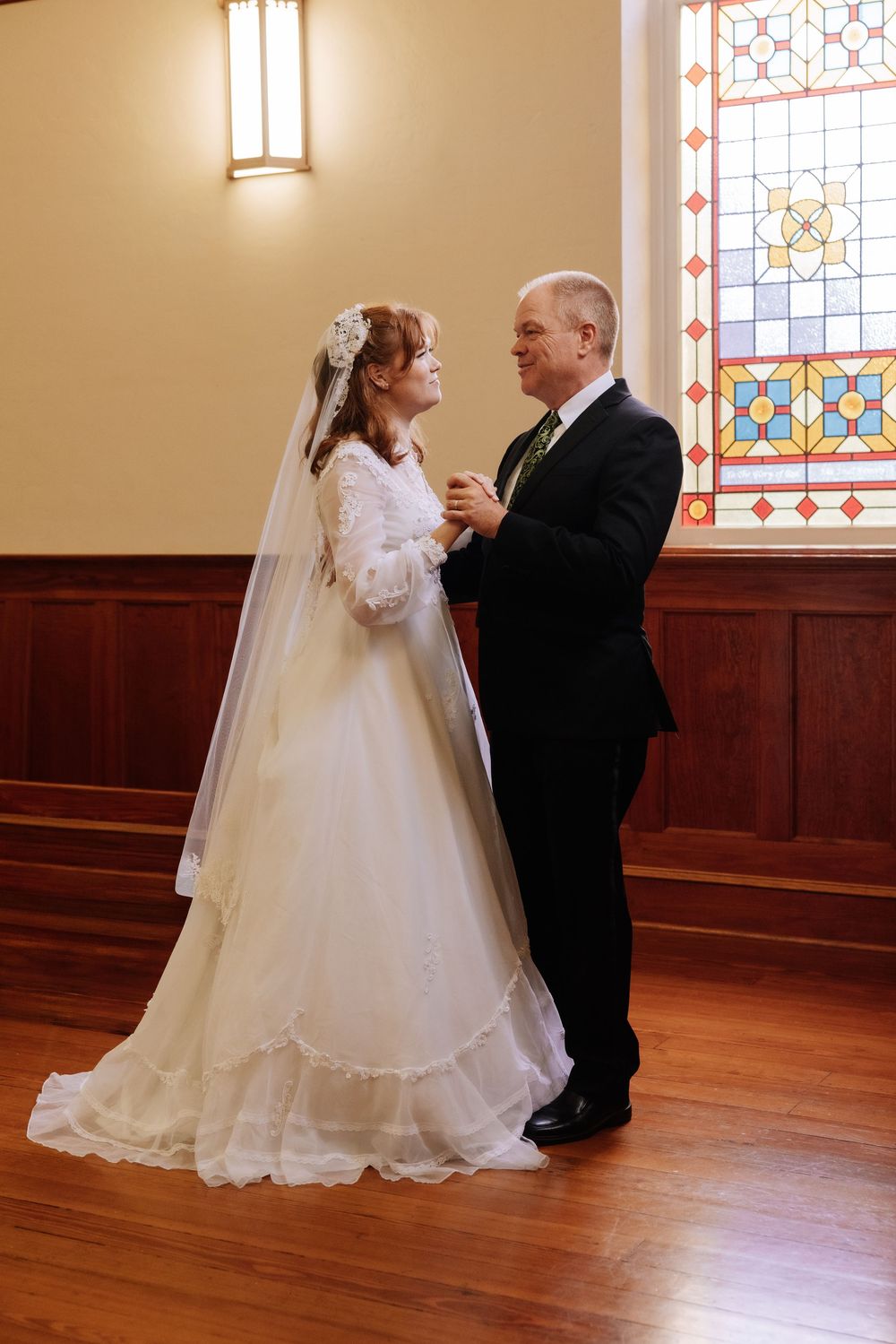 Bride and groom share a first dance near stained glass window in church, bride wearing vintage white gown with veil.