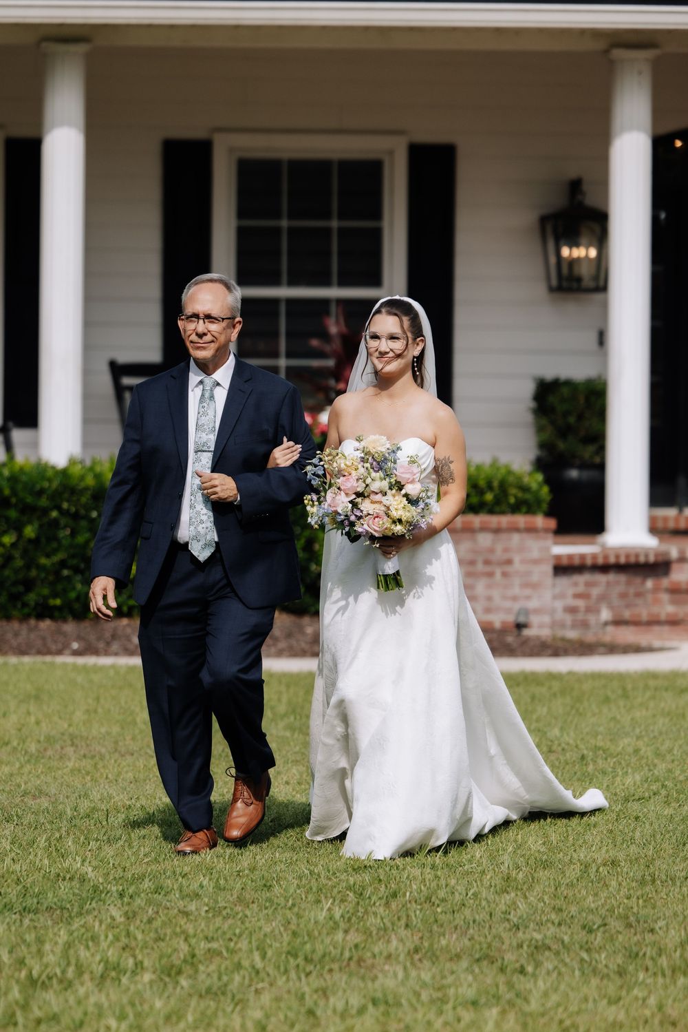 Bride in white gown holding floral bouquet walks with suited man across green lawn before white house.