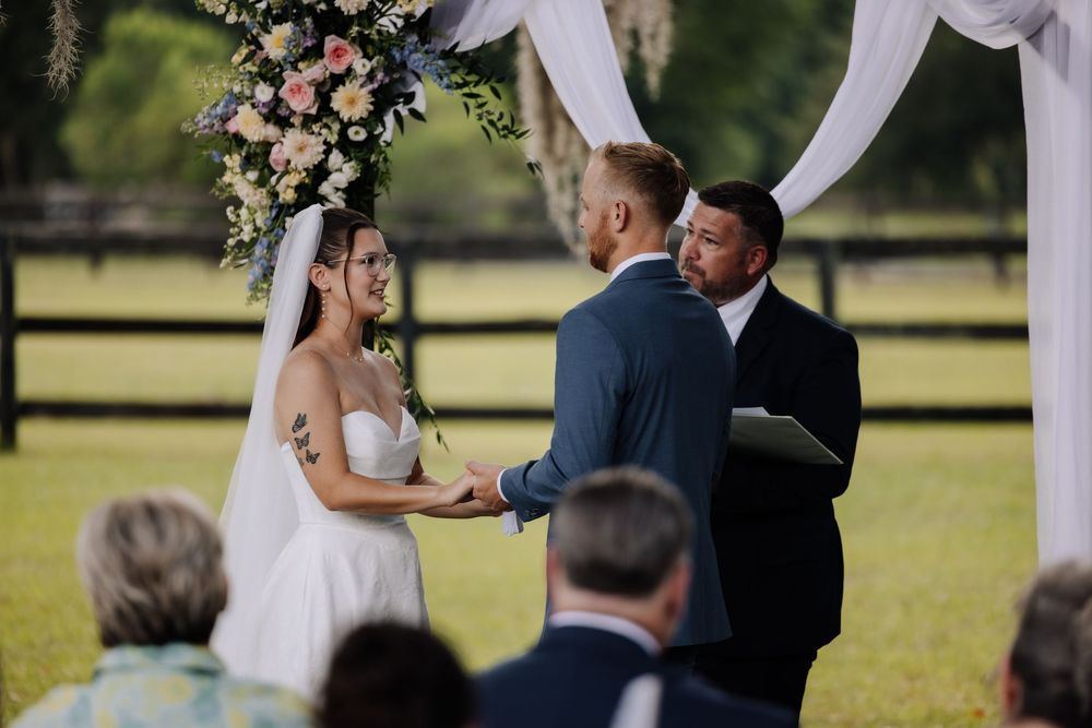 Bride and groom exchange vows at outdoor wedding ceremony beneath floral arch with white draping on green farm.