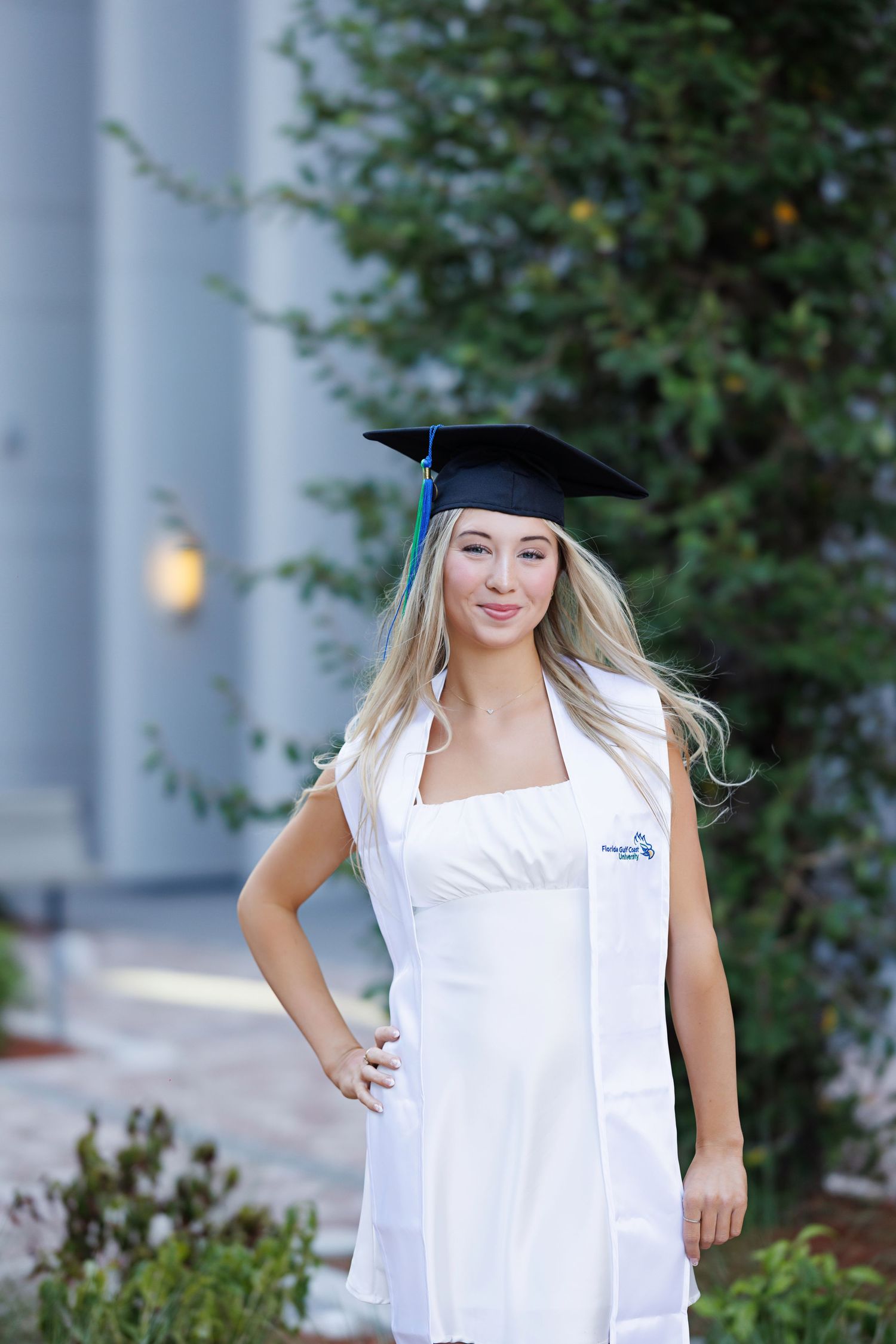 Graduate in white dress and cap poses outdoors near garden setting.