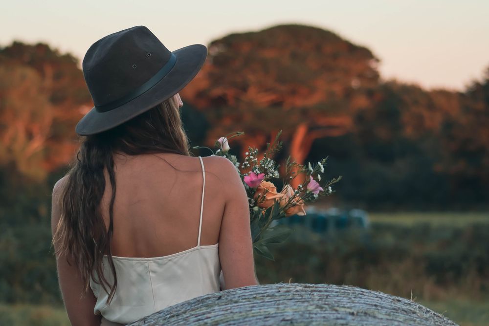 A person in a white dress and wide-brimmed hat stands in a golden field during sunset.