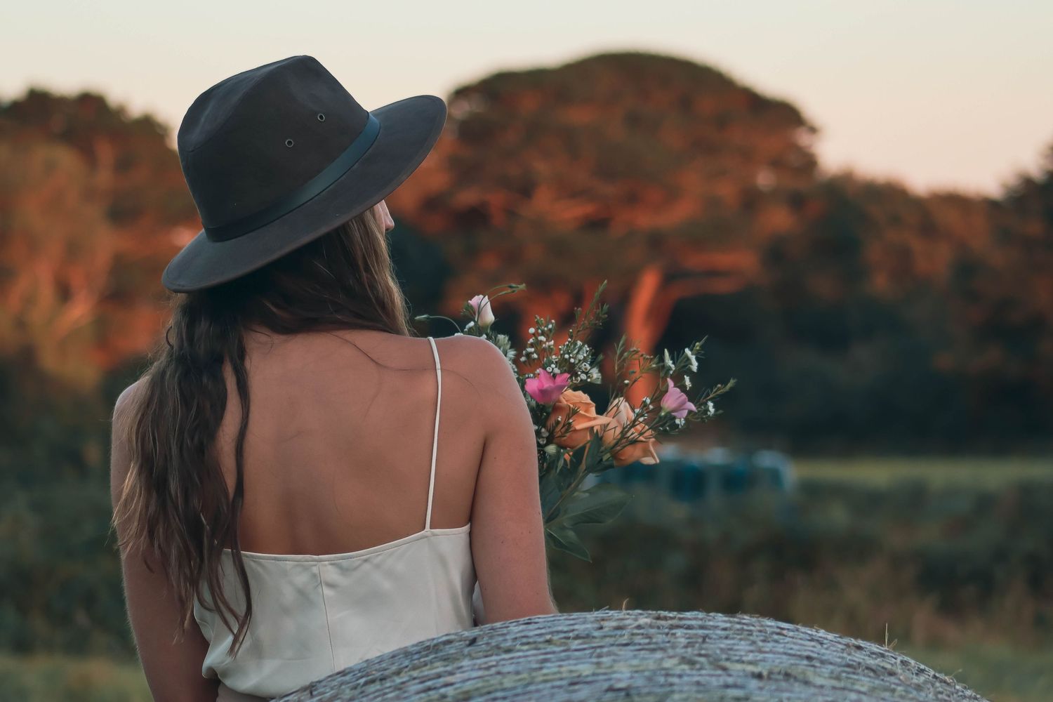 A person in a white dress and wide-brimmed hat stands in a golden field during sunset.