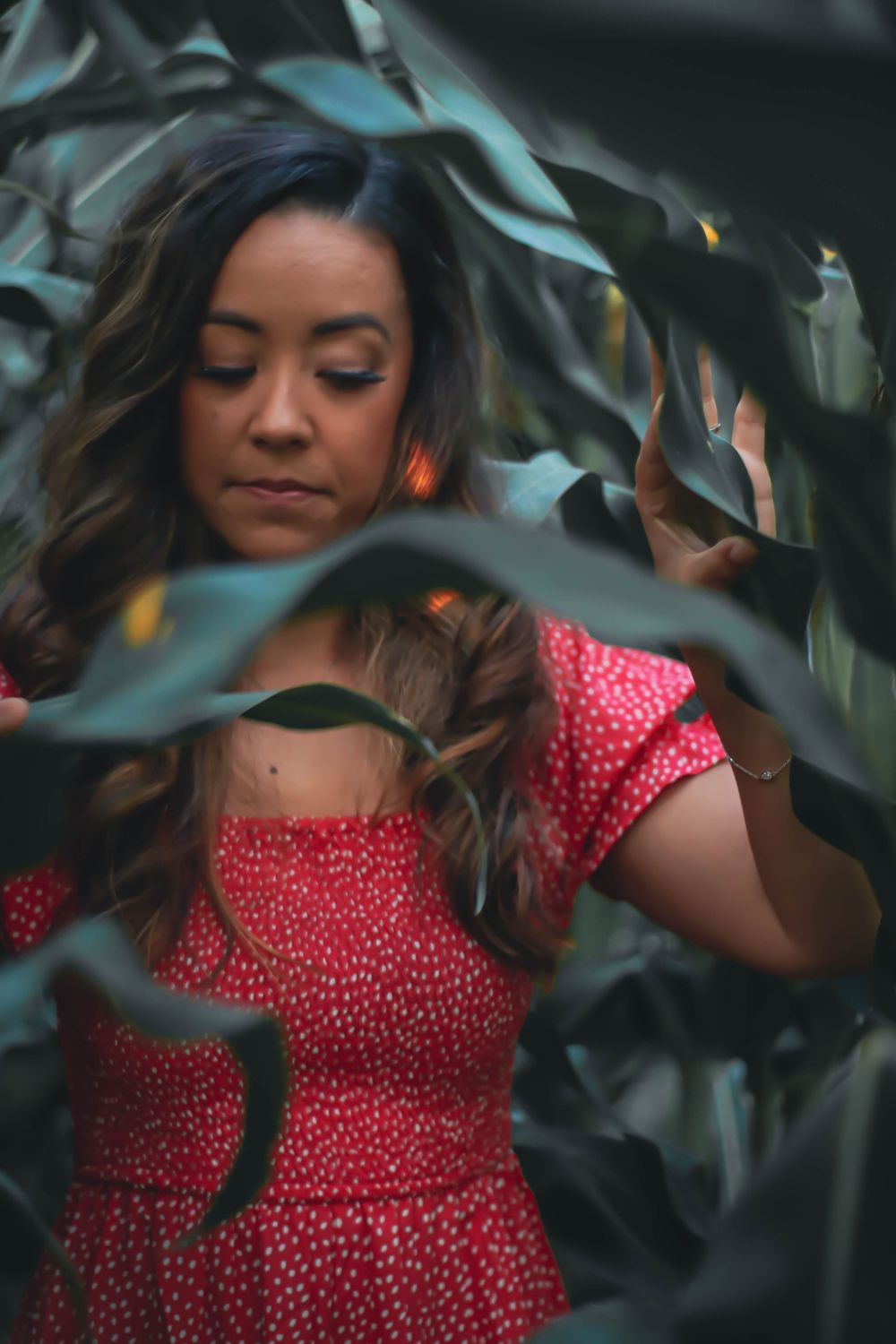 A person in a red polka dot dress poses thoughtfully among green foliage in a stylized portrait series.