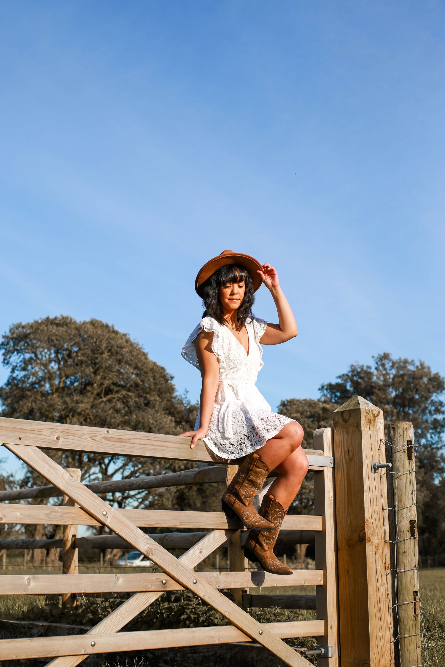 A dreamy country lifestyle portrait of someone in a white dress and boots sitting on a wooden gate.