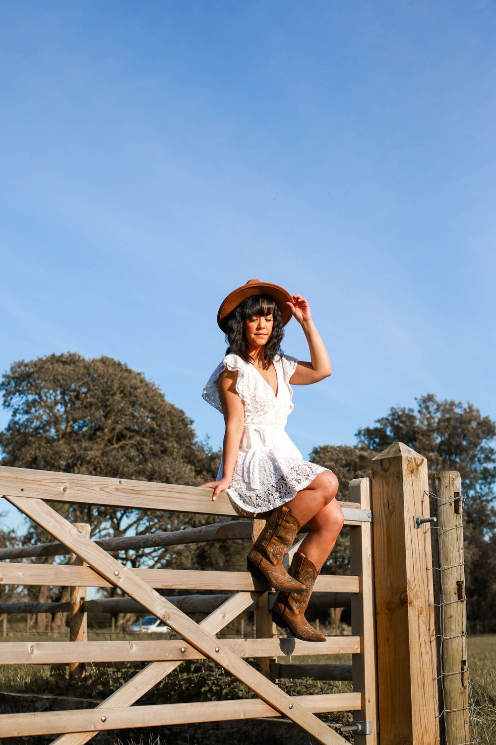 A dreamy country lifestyle portrait of someone in a white dress and boots sitting on a wooden gate.