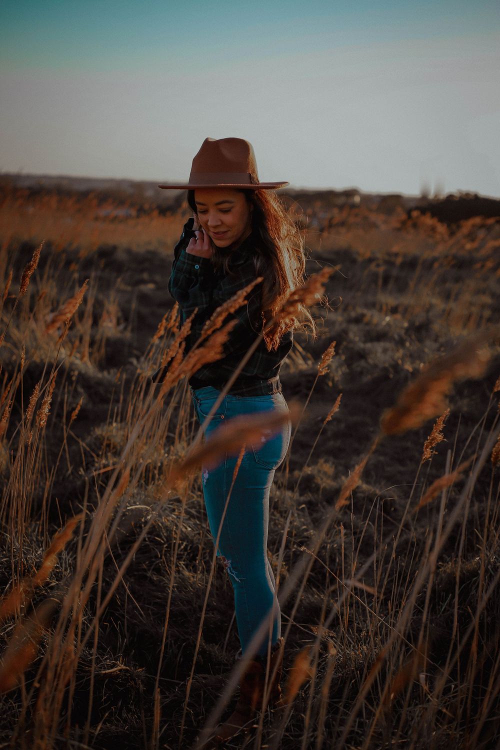 A person in a brown hat and denim stands in a golden field at sunset with tall grass surrounding them.