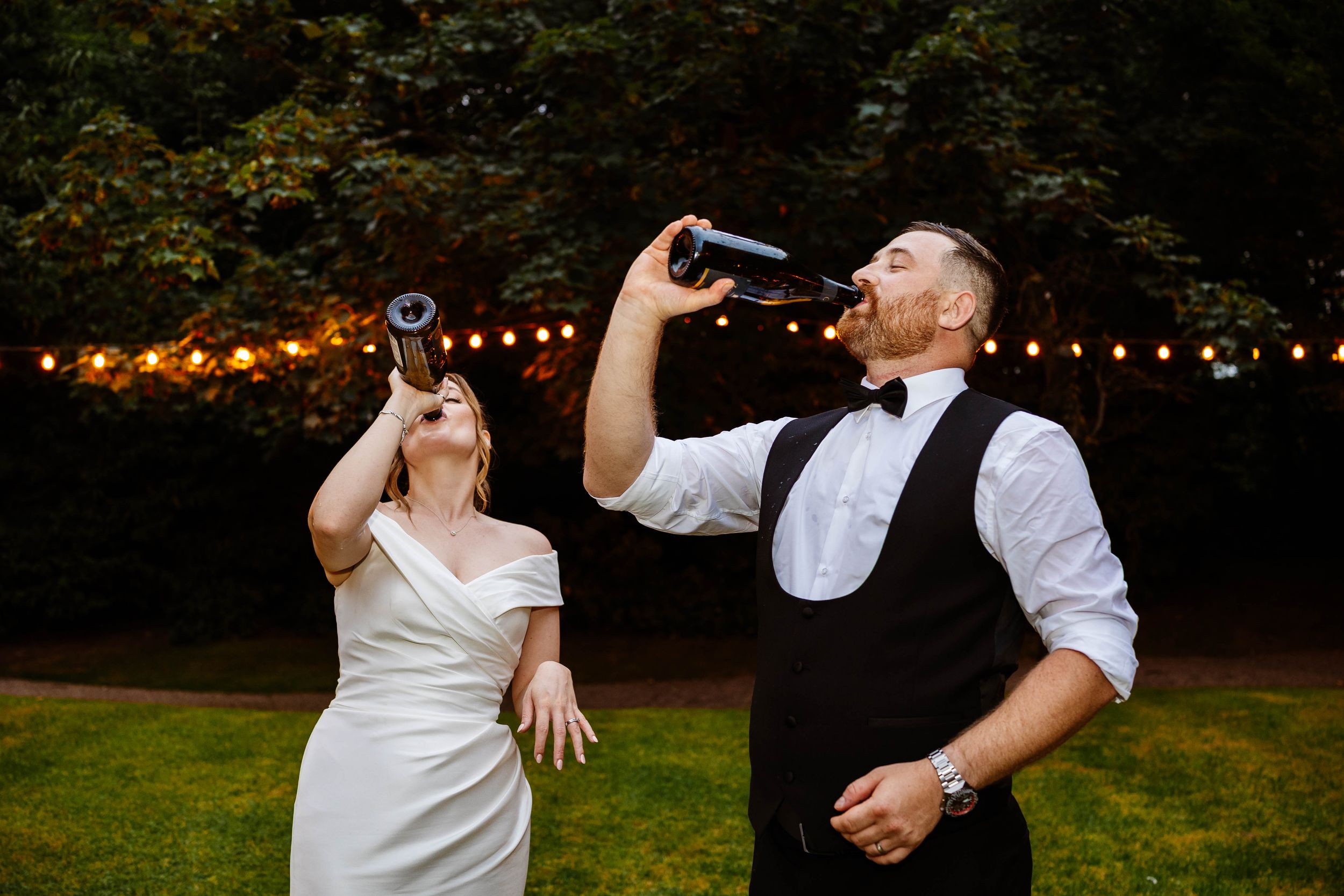 Bride and groom drinking from bottles outdoors at wedding reception with string lights in background at Bartle Hall, Preston