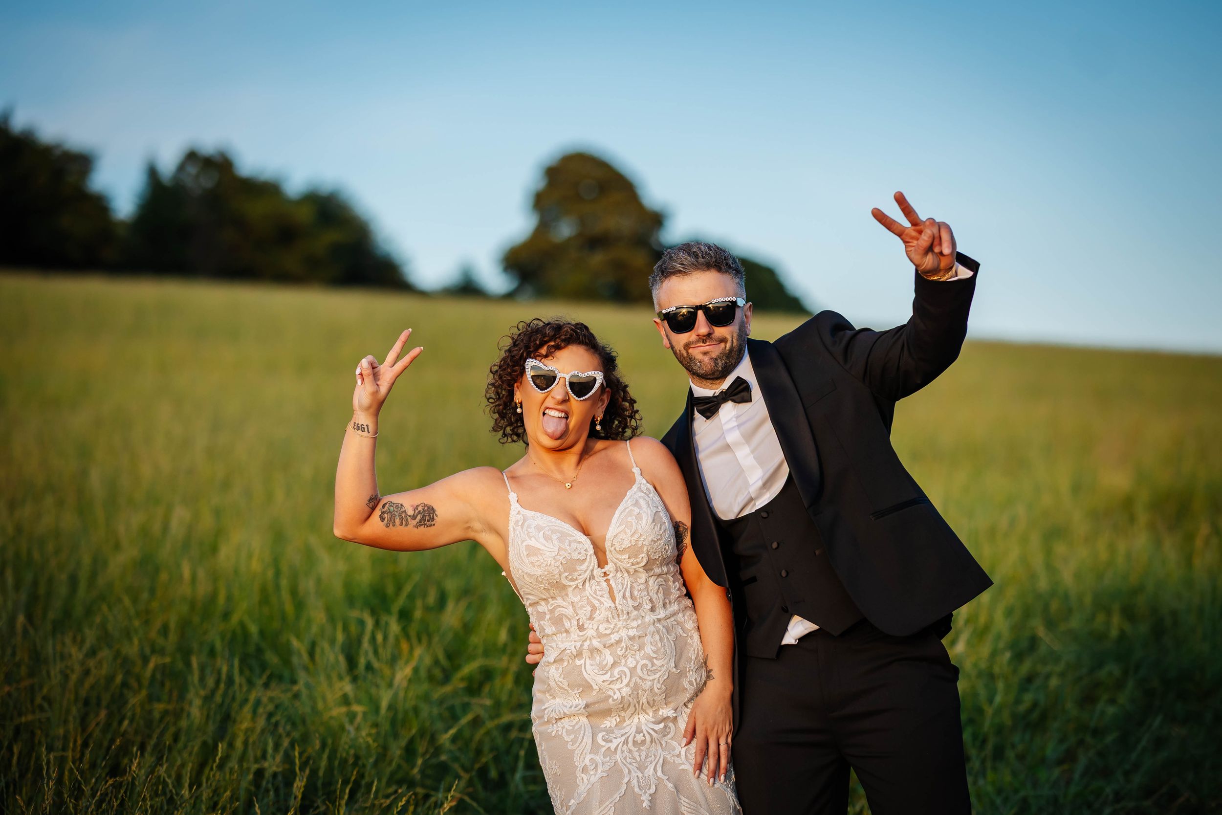Newlyweds in sunglasses flashing peace signs in a sunny green field, bride in lace gown & groom in tuxedo, Ribby Hall Wedding Photographer