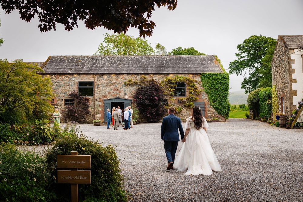 A couple walks towards a rustic stone barn building along a gravel path surrounded by lush countryside captured at Low Hall Wedding Venue