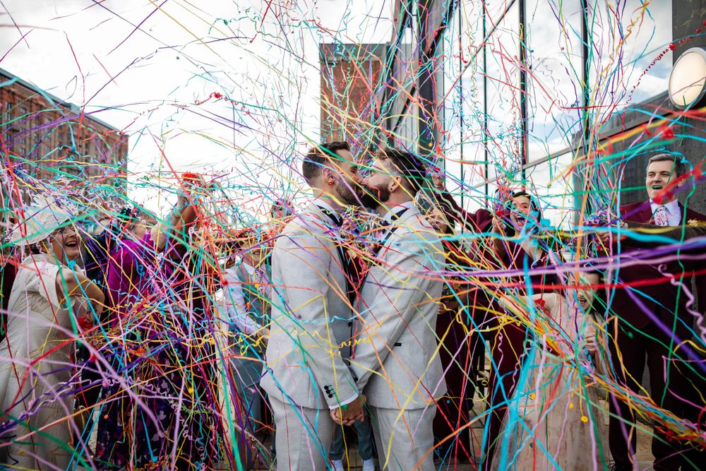 two grooms share a kiss during their confetti throw at the titanic hotel, Liverpool. guests are using paper streamers by Lynette Matthews