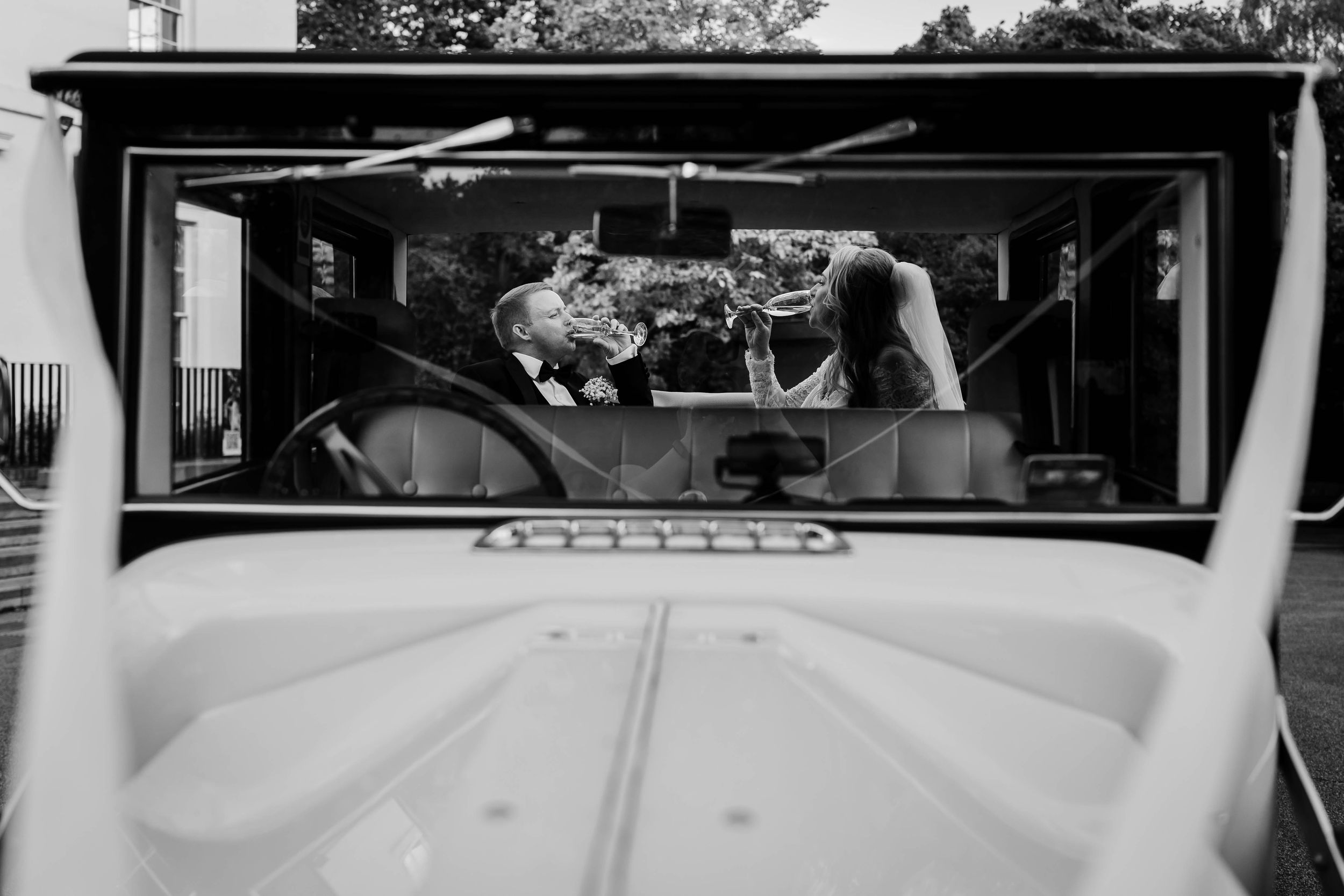 Black & white photo of a bride & groom drinking champagne inside a wedding car, seen through the windshield by Lynette Matthews Photography