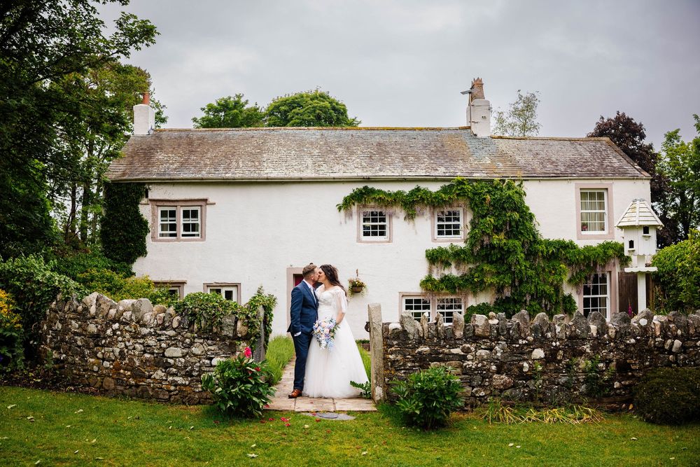Two people share an intimate moment in front of a charming white countryside cottage with a stone wall and lush gardens - Low Hall Weddings