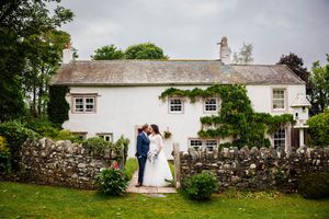 Two people share an intimate moment in front of a charming white countryside cottage with a stone wall and lush gardens - Low Hall Weddings