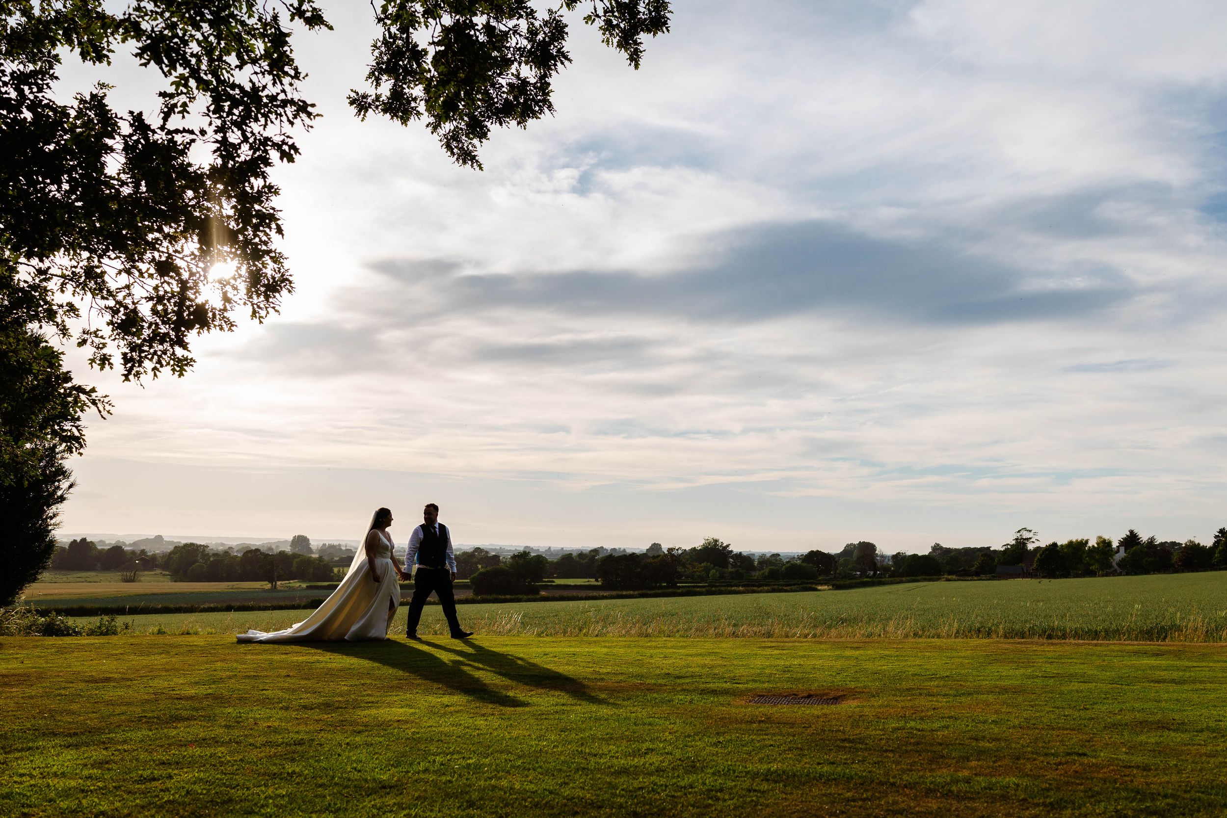Bride and groom walking hand in hand across a lush green field at sunset on their wedding day at West Tower Exclusive Wedding Venue