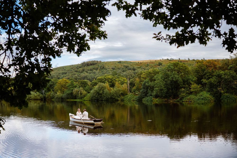 A bride & Groom rowing a small boat on a calm lake surrounded by trees and rolling hills at Wyresdale Park captured by Lynette Matthews