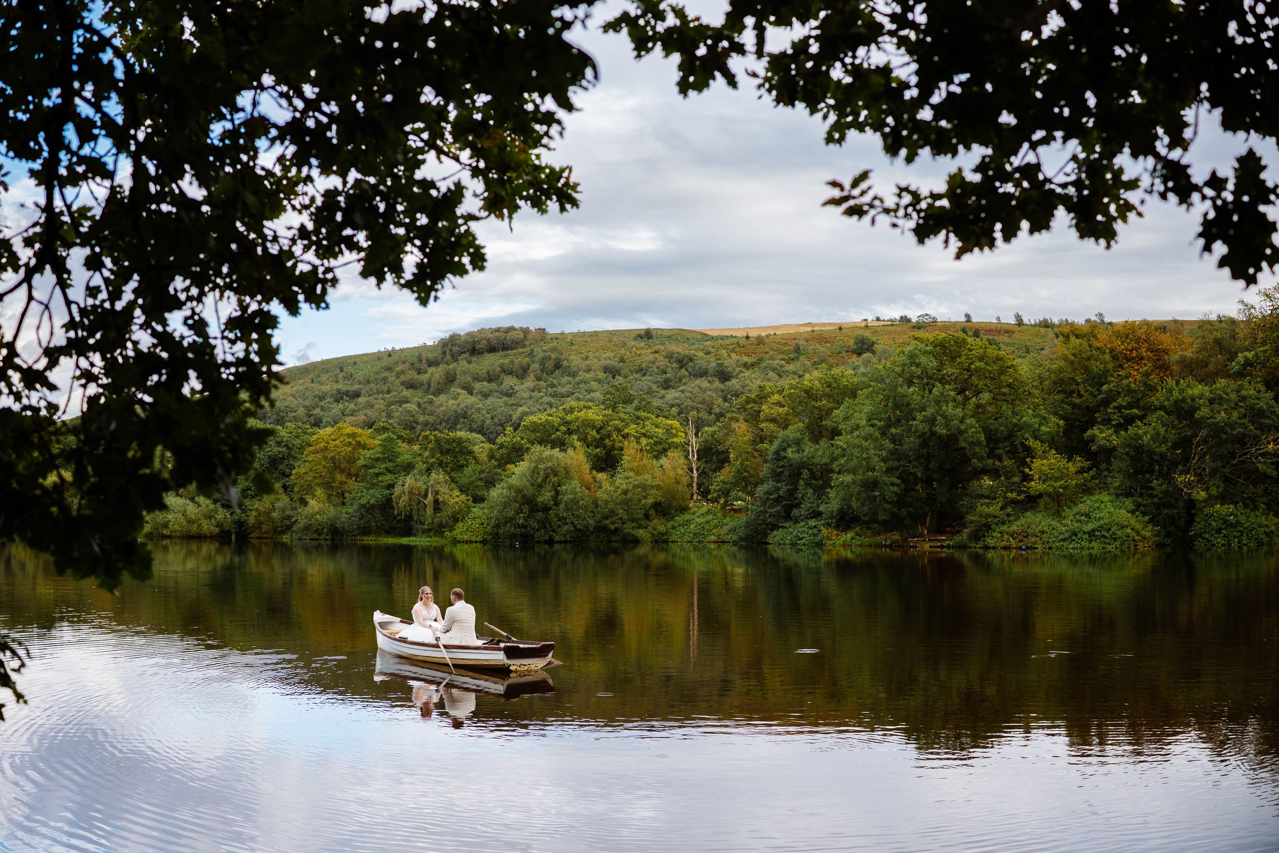 A bride & Groom rowing a small boat on a calm lake surrounded by trees and rolling hills at Wyresdale Park captured by Lynette Matthews